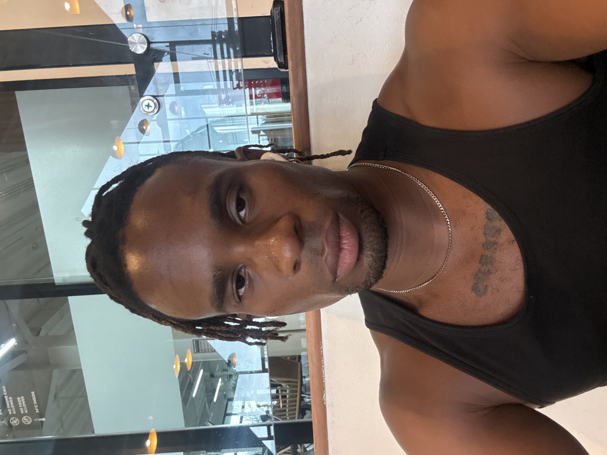A medium indoor shot, taken during daylight, depicts a man in a black tank top. He has dark skin, short braided hair, and a light beard. A silver chain is visible around his neck, and a tattoo is located on his left pectoral area. The man is looking directly at the camera. The background features a window, reflecting a bright interior with hanging lights and what appears to be a cafe setting with seating. The location is within the city of Unknown, Unknown.