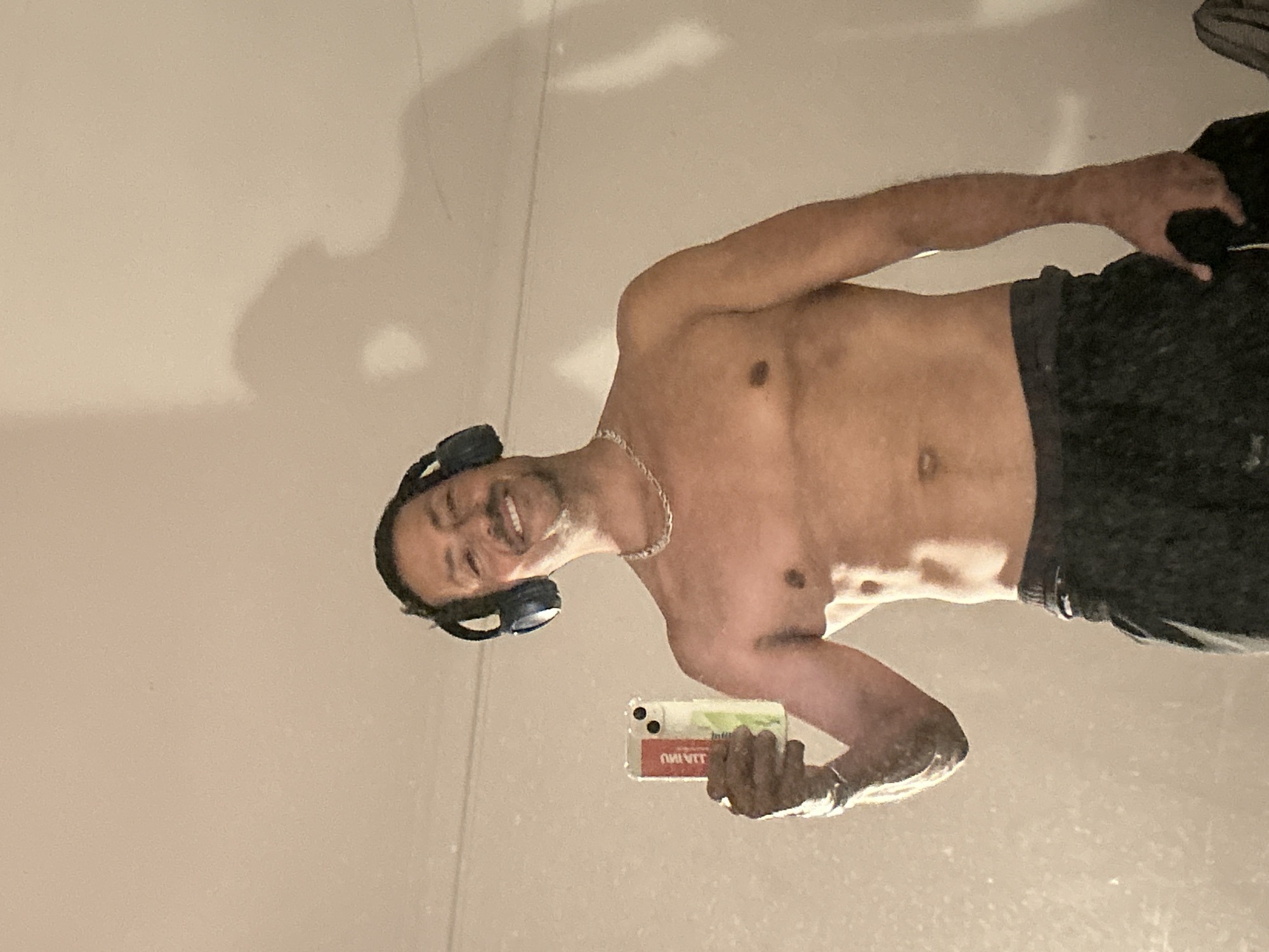 A man with dark hair, a mustache, and a goatee is taking a mirror selfie. He is wearing dark headphones and a silver chain. He is holding a smartphone in his left hand. The man is shirtless, showing his chest and abdomen. He is wearing dark sweatpants. The background is a plain, light-colored wall. There is a shadow of the man on the wall behind him. The scene appears to be indoors. There is no information to indicate the location is in Barcelona, Spain.