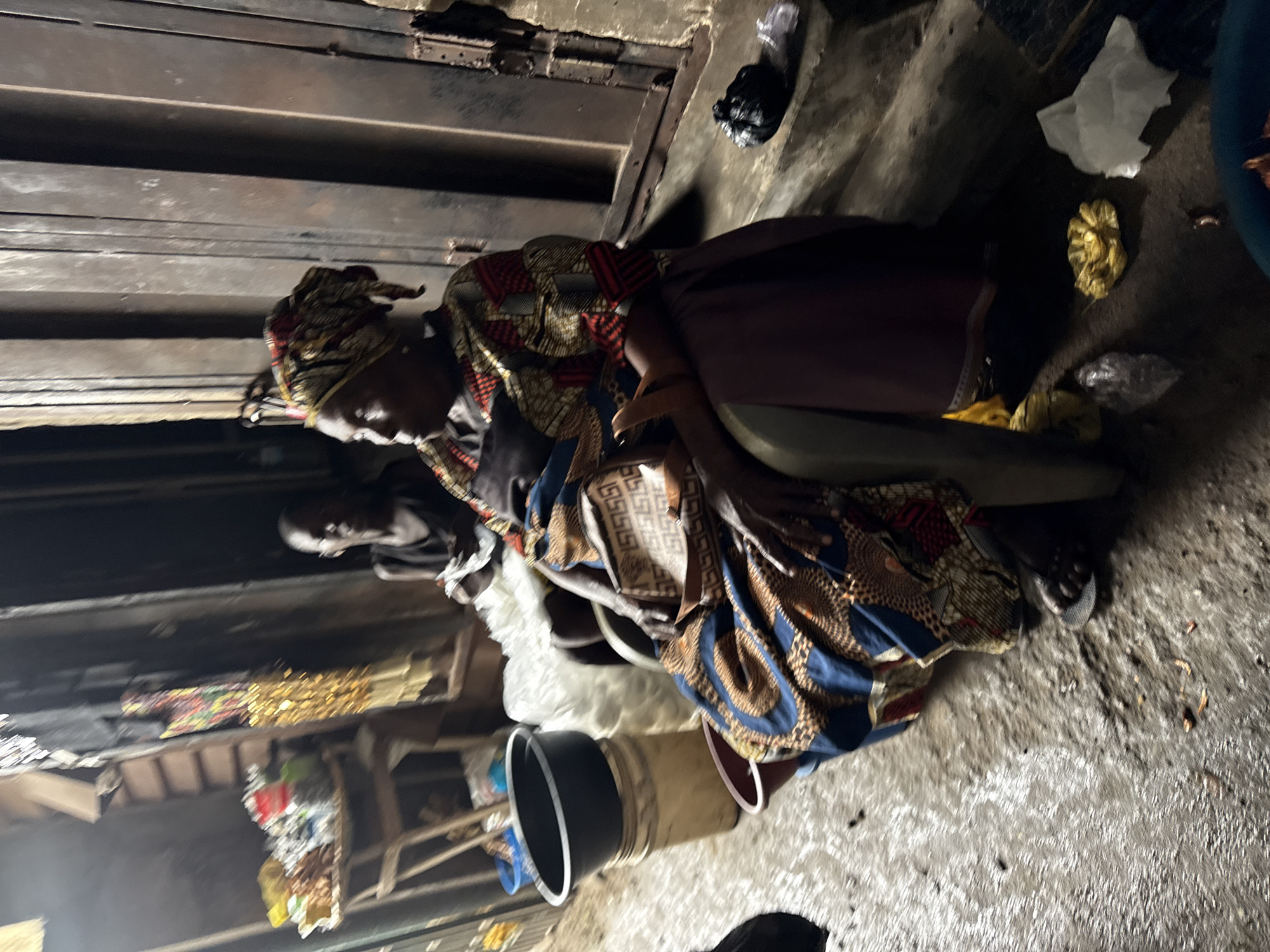 The image depicts a scene from a Lagos, Nigeria market, likely an outdoor or semi-enclosed stall area. Two individuals are present. In the foreground, an adult woman is seated in a chair, facing left, and wearing a headscarf and a vibrantly patterned traditional garment. Her arms are resting on her lap, and she is holding a small, patterned handbag. To her right, a younger individual, possibly a child, is also seated, looking towards the left.
The environment appears to be a busy market setting. Various objects are scattered around, including plastic buckets, a wooden stand with items on display (possibly goods for sale), and discarded plastic bags and wrappers on the ground. The ground surface is uneven and appears wet or damp in places, suggesting it may have rained or there's a water source nearby. The background consists of what looks like corrugated metal sheeting, typical of market structures in Lagos. The lighting is dim, with shadows casting across the scene, indicating it could be late afternoon or early evening.