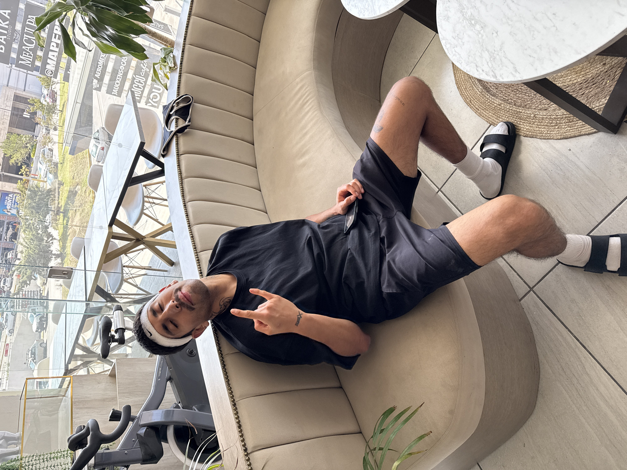 A young man is captured lounging casually on a curved, light beige upholstered sofa in a bright, modern interior, likely a cafe or a lounge area in Puebla, Mexico. He is lying on his back with his head propped up, looking directly at the camera while making a peace sign with his left hand. He wears a black t-shirt, black shorts, white socks, and black slide sandals. A white headband with a black swoosh logo (Nike) is visible on his forehead. Tattoos adorn his left forearm, displaying text that appears to read "REALME" or similar, along with a cross symbol, and small text on his right thigh, possibly "la te cafe". A cell phone or similar device is held in his right hand.
The setting is characterized by large glass windows that showcase a sunny daytime view of a city street, complete with parked cars and trees. Numerous reflections on the window reveal various business names and words, including "BAIKA STUDIO," "MAPFRE," "AEROMEXICO," "JAUCKENS CAS," "MUCCI café," "YOU," and Spanish terms like "SALE" and "RENTA."
Inside, the space features two small, round tables with white marble-patterned tops and dark bases, one placed on a woven placemat. A pair of black slide sandals rests on the sofa near the window. A black exercise spin bike is partially visible in the foreground, suggesting a connection to a fitness center or a wellness-oriented space. The flooring consists of light-colored tiles, and potted green plants add natural decor to the clean, contemporary environment. The man's relaxed demeanor and peace sign convey a comfortable and pleasant mood.