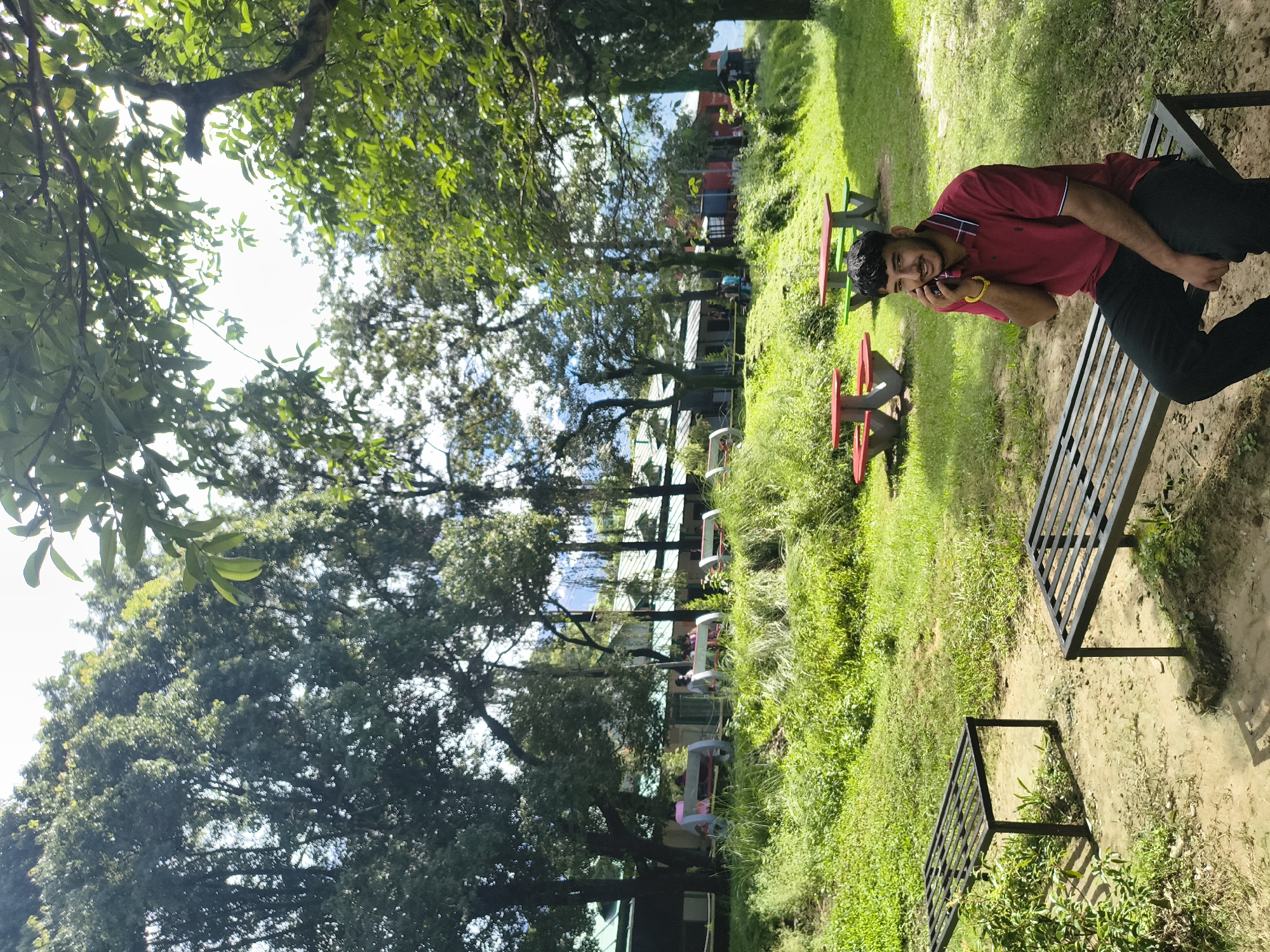 A young man with dark hair and a beard, wearing a red polo shirt and black pants, is sitting on a metal bench outdoors. He is holding a cell phone to his right ear and appears to be in conversation. He is looking towards the camera with a slight smile. The bench is situated on a dirt path in a grassy, park-like environment with several trees. In the background, there are more trees, buildings with green roofs, and other park amenities like picnic tables and benches. The overall scene suggests a relaxed outdoor setting in Dharamshala, India, likely a public park or a campus green space. The lighting indicates it is daytime, with sunlight filtering through the trees.