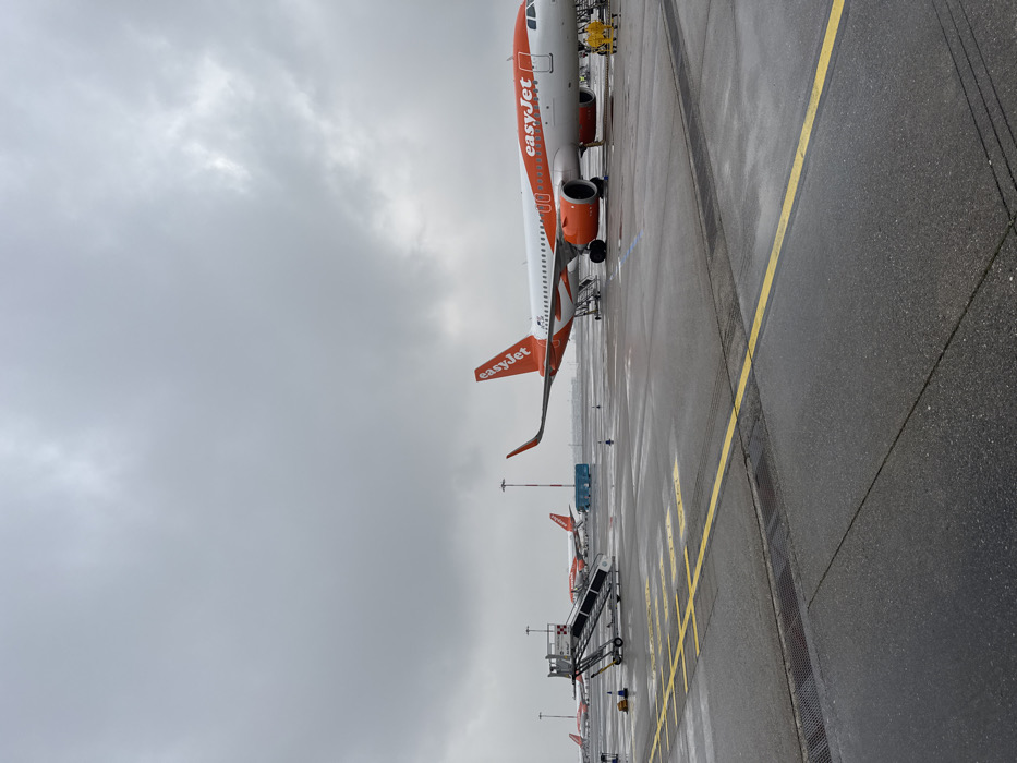The scene depicts an airport tarmac at Milan Malpensa Airport in Somma Lombardo, Italy, under an overcast, uniformly gray sky. The asphalt surface is wet with standing water and reflections, consistent with recent precipitation.
In the mid-ground, a white and orange EasyJet Airbus A320 series aircraft is parked, positioned diagonally with its nose facing towards the left. Passenger boarding stairs are deployed and connected to both the forward and aft main passenger doors. Two individuals wearing yellow high-visibility clothing are visible on the tarmac near the aircraft's front landing gear.
To the left foreground, a standalone silver mobile passenger stair unit, featuring red and white checkered safety markings on its top platform, rests on the wet tarmac. Multiple other EasyJet aircraft in identical livery are visible parked in the distance, indicating an active airport apron. Yellow operational markings are painted on the tarmac surface, defining various areas. Tall utility poles are scattered across the apron.
