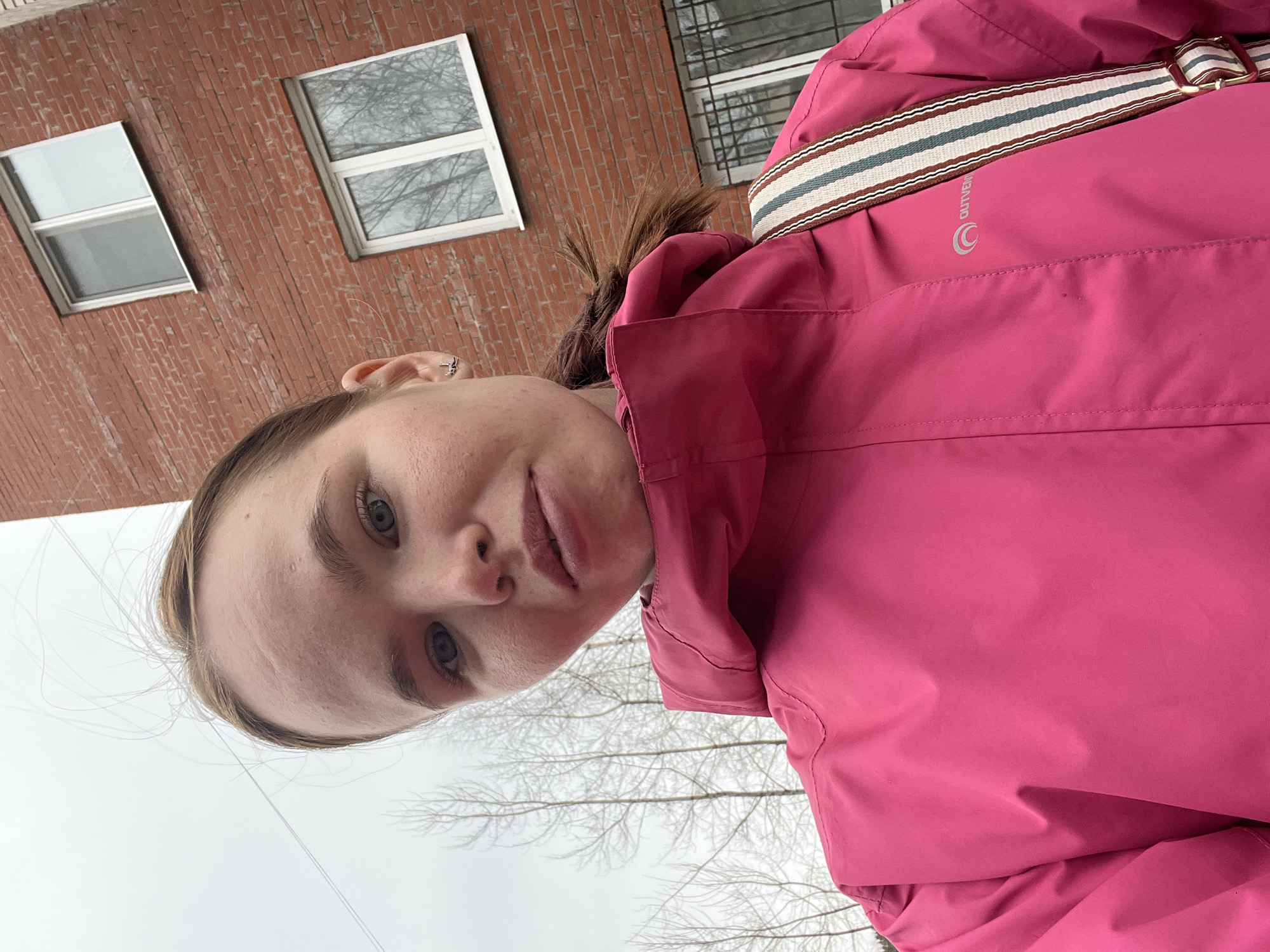 A young woman with fair skin and blue eyes, wearing a bright pink winter jacket with a striped strap across her shoulder, is taking a selfie outdoors. A red brick building with white-framed windows is visible in the background. Bare tree branches are also visible. The location is most likely in the city of Chaikovsky, Russia, based on the prompt.