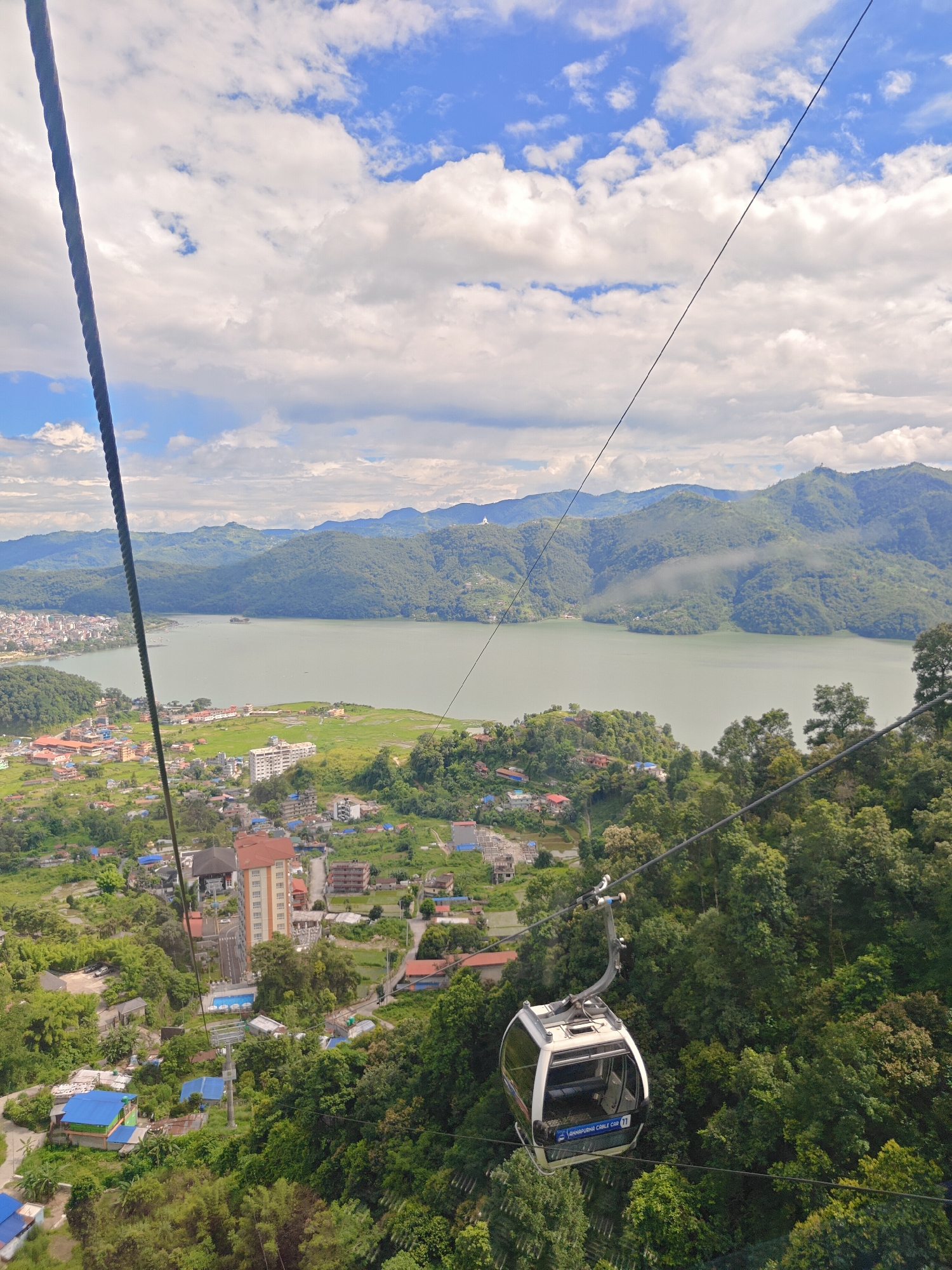 The image captures a view from a cable car in Pokhara, Nepal. The cable car itself is a modern, white cabin with large windows, suspended from a thick cable. Below, a lush green landscape unfolds, dotted with houses and buildings, many featuring blue rooftops. A prominent orange-tinted building stands out among the residential structures. In the distance, a wide, calm lake, likely Phewa Lake, stretches out, reflecting the partly cloudy sky. Rolling, verdant hills form the backdrop, with a hazy atmosphere obscuring the farthest peaks. The sky is a vibrant blue, interspersed with fluffy white clouds, suggesting fair weather. The scene conveys a sense of elevation and a panoramic perspective of the city and its natural surroundings.