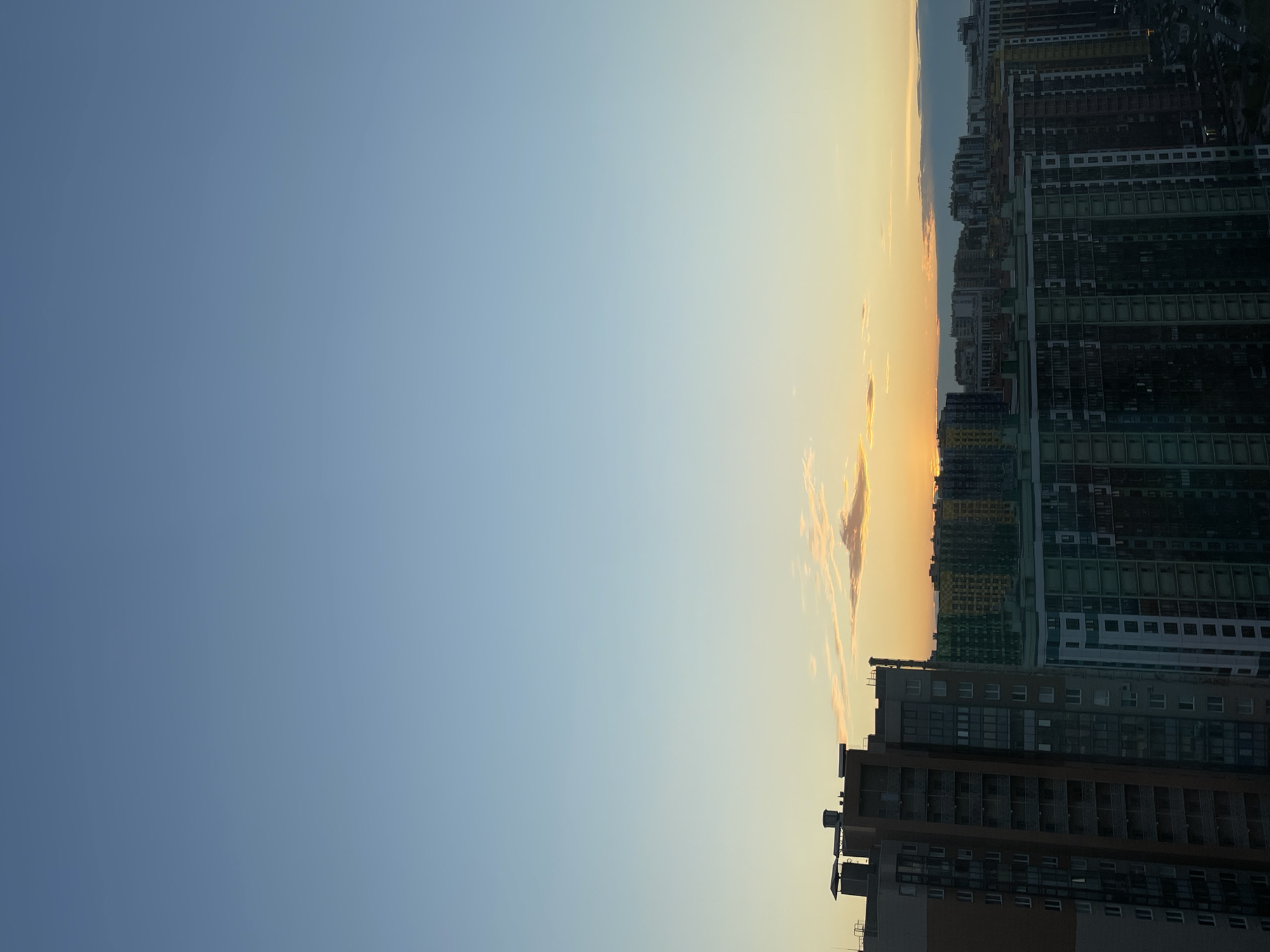 This vertical, wide shot captures a sunset over a densely populated residential area in Saint Petersburg, Russia. The sky is a gradient of clear blue near the top, transitioning to pale yellow and orange near the horizon, indicative of the sun setting. A few thin, wispy clouds are visible, tinged with the colors of the sunset.
In the foreground, the upper floors of several multi-story apartment buildings dominate the lower half of the frame. The building on the left is predominantly brown and grey, with visible windows and balconies. To its right, a row of taller apartment buildings with colorful facades in shades of green, yellow, and blue are prominent, showcasing their repetitive window patterns. Further in the background, more apartment buildings of varying heights and architectural styles form a dense urban landscape, stretching towards the horizon. The overall impression is of a modern, large-scale housing development.
There are no people, animals, or vehicles clearly visible in the frame. The scene is a still, observational shot of the urban environment bathed in the warm light of the setting sun. The time of day is clearly evening, as suggested by the sunset and the diminishing light on the buildings. The environment is purely urban, characterized by the presence of numerous residential buildings.