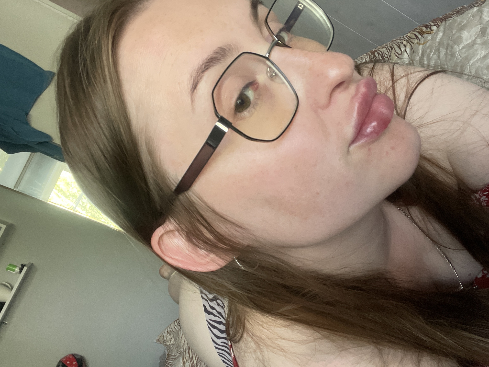 The image is a close-up, low-angle selfie of a young Caucasian woman with long brown hair, wearing glasses and a silver necklace. She is lying down on a bed with a patterned comforter. Her face is turned towards the camera, and she is looking slightly upwards. The background shows a wall with a shelf and a window with natural light streaming in. The scene appears to be indoors, likely in a bedroom. There is no information in the image that would suggest a location in the city of Сырково, Russia.