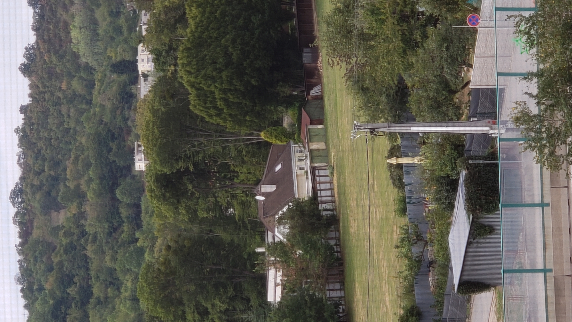 The image is a medium-distance, vertical shot of a residential area in Prešov, Slovakia. The foreground shows a concrete barrier and a green-railed fence, beyond which a corrugated metal shed is visible, partially overgrown with ivy. Further back, a paved area leads to a fence bordering a large, grassy field. Behind the field, a house with a brown roof and a white dormer window sits in front of a line of garages with green doors. The house is surrounded by trees and a wooden fence. In the background, a densely wooded hillside rises, with a path visible on the upper slope. The sky is overcast. The image appears to be taken on a cloudy day, possibly in late summer or early autumn, given the lush green foliage.
