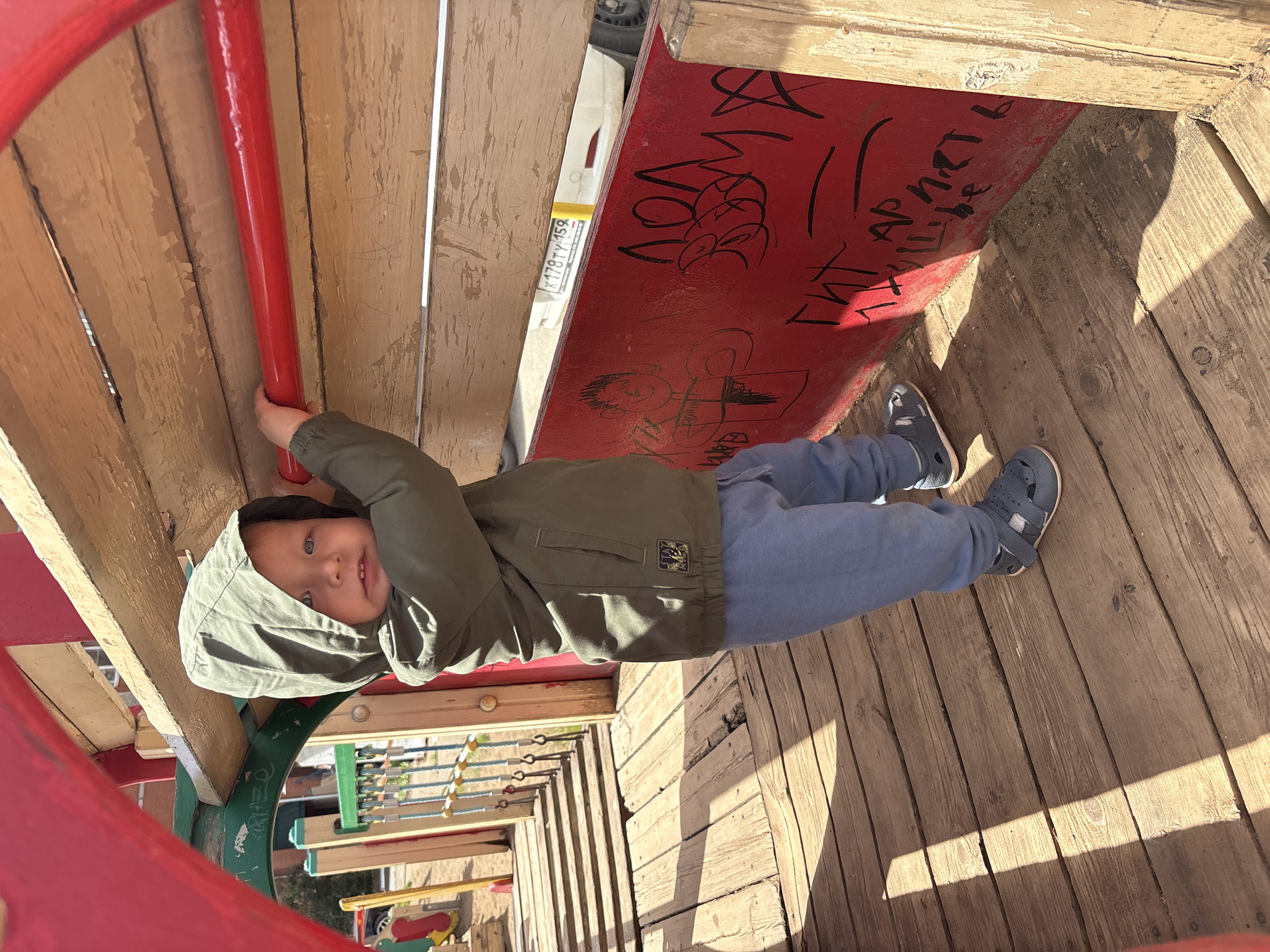 A young child stands on a wooden walkway of a playground structure. The child is wearing a green hooded jacket, blue pants, and grey sandals. They are holding onto a red horizontal bar. The walkway is made of weathered wooden planks and is illuminated by sunlight casting shadows. In the background, there is playground equipment and a white car with a license plate. Red graffiti is visible on a red panel on the right side of the image. The location is the city of Chaikovsky, Russia.