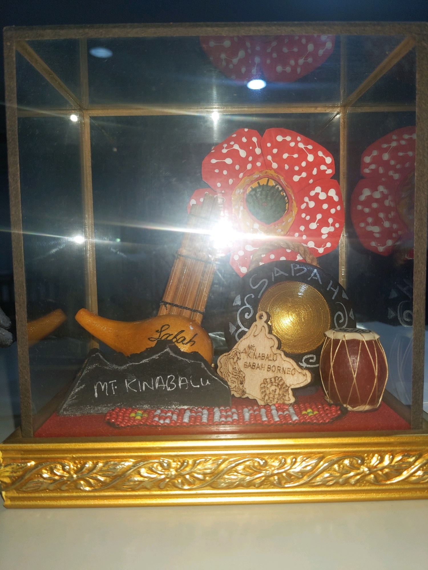 A diorama is displayed within a glass-fronted case. The case has a golden ornate base and a wooden frame. The diorama features miniature representations of several items. Prominently displayed are two large, red flowers with white spots, resembling Rafflesia flowers. In the foreground, a miniature mountain labeled "MT. KINABALU" is depicted. To the left of the mountain is an orange gourd-like object with "Sabah" written on it. Behind the mountain, there is a small drum and a circular object that appears to be a gong. A wooden keychain with an outline of Sabah and text is also visible. The background is dark and reflective, showing the interior of the display case. The scene appears to be a souvenir or cultural display, potentially located in Kolombong, Malaysia, given the references to Sabah and Mount Kinabalu. No people are present in the image.