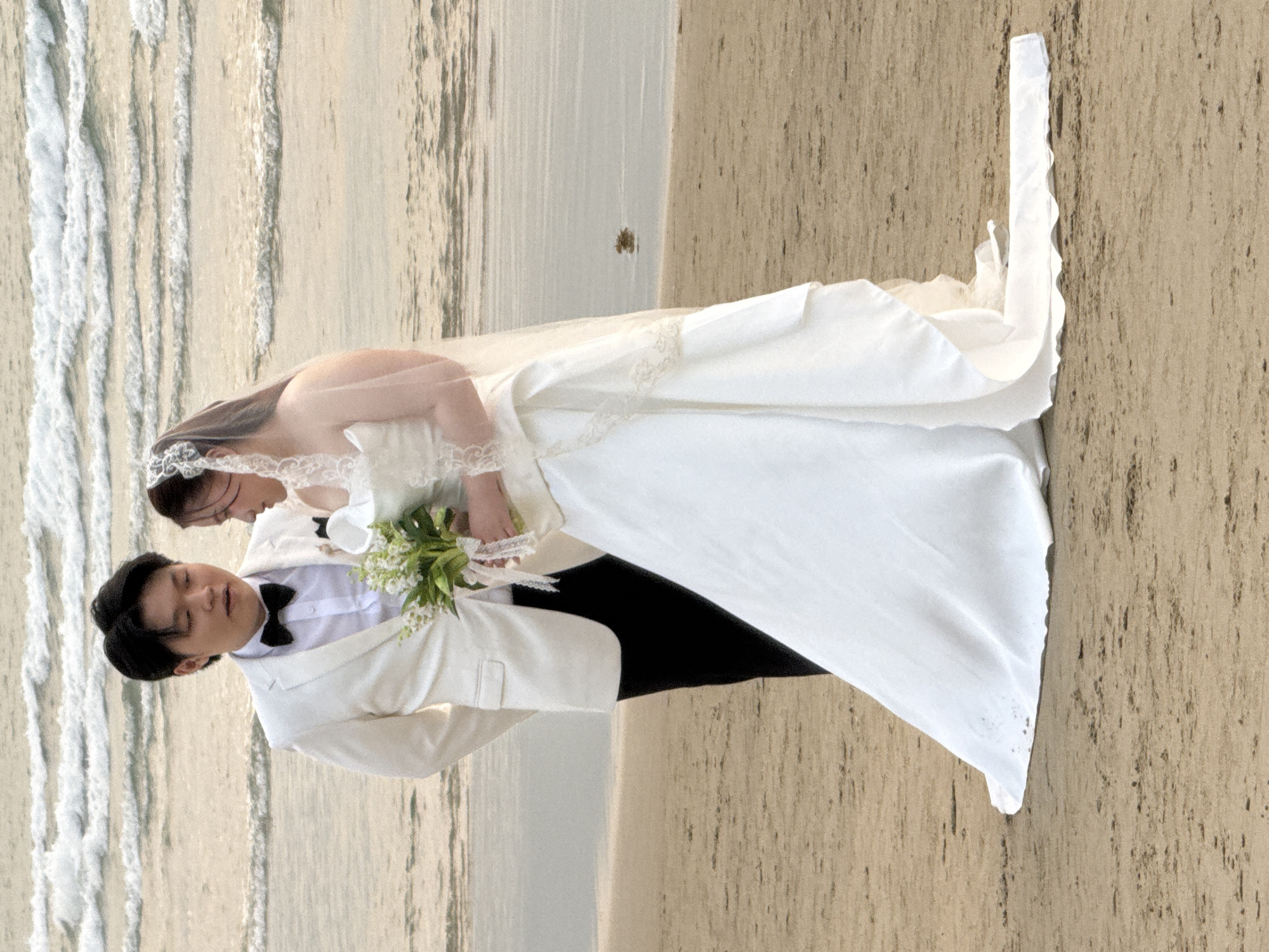A wedding photo captures a couple on a beach. The bride wears a white wedding dress and veil, holding a bouquet. The groom is in a white suit with a black bow tie. The scene appears to be near the city of Đà Nẵng, Vietnam, as indicated by the beach and ocean environment. The couple stands on the sand with the sea visible in the background.