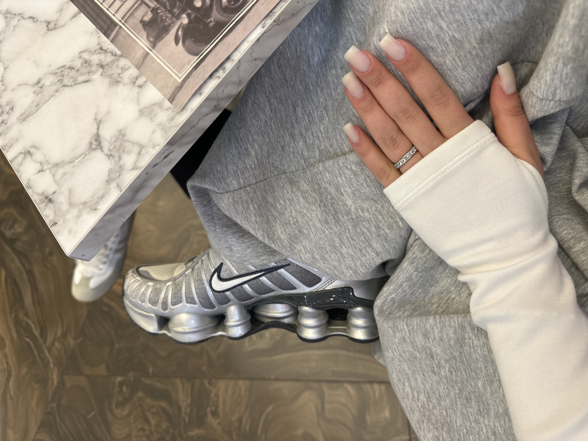 The image offers a close-up, top-down perspective of a person's lower body and left hand in an indoor setting in Yerevan, Armenia. The subject is wearing comfortable, casual attire, including light gray sweatpants or joggers and a white long-sleeved top with thumbholes. On their left foot, a distinctive silver and grey Nike Shox sneaker is visible, featuring a black Nike swoosh and the signature column cushioning.
The person's left hand rests gently on their leg, showcasing manicured, square-shaped fingernails in a milky white or nude tone. A delicate ring, adorned with a line of small, sparkling stones, is worn on the ring finger.
To the upper left of the frame, a corner of a white table with a prominent gray marble pattern is visible. A book or magazine rests on this table, open to a page displaying a black and white image, which appears to be of a classic vehicle like a car or motorcycle, framed by white lines. The text on the page is not legible. The floor beneath the subject features a polished surface with swirling patterns in shades of brown and beige, resembling natural stone or decorative tiling.
The scene conveys a sense of casual comfort and relaxation. The lighting is even, suggesting an indoor environment during daytime or a well-lit evening. No specific activity is captured beyond the person resting their hand and foot.