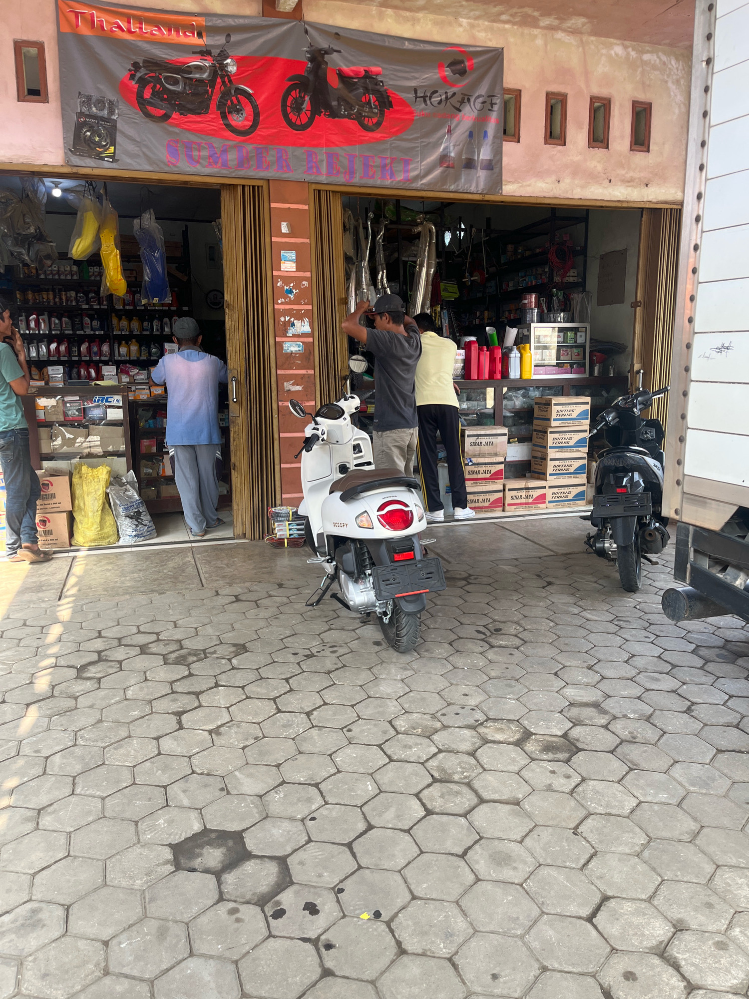 In Kemuning, Indonesia, the exterior of a storefront is captured. A banner with images of motorcycles and the text "THAIZANO HOKAGE SUMBER REJEKI" stretches across the top. Below, the open entrance reveals shelves stocked with various products, including bottles of oil and what appear to be automotive parts. Three men are present: one in a green shirt and jeans stands near the entrance, another in a blue shirt and grey pants is inside the shop, and a third in a grey shirt and dark pants is near the doorway. Two motorcycles are parked outside: a white scooter on the left and a black motorcycle on the right. The ground is paved with hexagonal gray tiles, with some oil stains visible. The storefront appears to be an auto parts or repair shop.