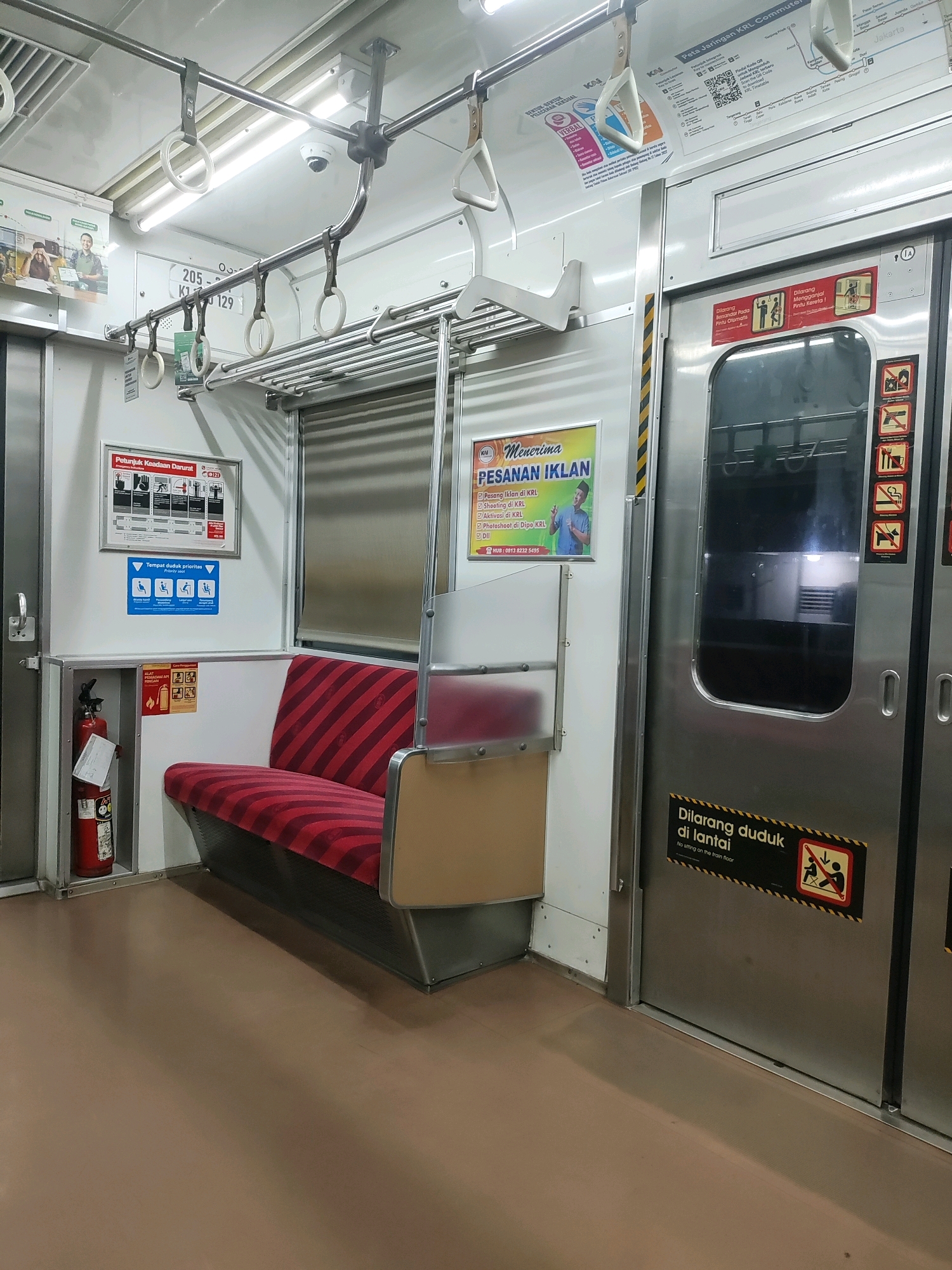 The image depicts the interior of a train carriage, likely within the city of Tambun, Indonesia. The scene is oriented towards the right side of the carriage, showcasing a section of seats and a train door.
On the left, a red and white striped bench seat is visible, positioned against a white wall. To the left of the seat is a red fire extinguisher mounted in a metal compartment. Above the seat, various informational signs are affixed to the wall, including one detailing emergency procedures with pictograms and another indicating priority seating for specific groups.
The dominant feature on the right is a large, metallic sliding train door, likely leading to another carriage or the exterior. A yellow and black striped sticker is affixed to the door, bearing the text "Dilarang duduk di lantai" which translates to "No sitting on the train floor," accompanied by a pictogram of a person sitting on the floor with a red cross over it. Above this, a smaller red sign with pictograms illustrates rules regarding door usage.
Further to the left, a yellow poster is mounted, advertising "Menerima Pesanan Iklan" (Accepting Ad Orders), detailing services such as ad placement, shooting, and photoshoots within the KRL (Kereta Rel Listrik, electric rail train) network.
The carriage interior is illuminated by overhead fluorescent lights. Several metal handrails and grab handles are visible along the ceiling and above the seats, typical for public transport. The floor appears to be a plain, light brown material. The overall impression is of a functional, well-maintained public transportation environment.