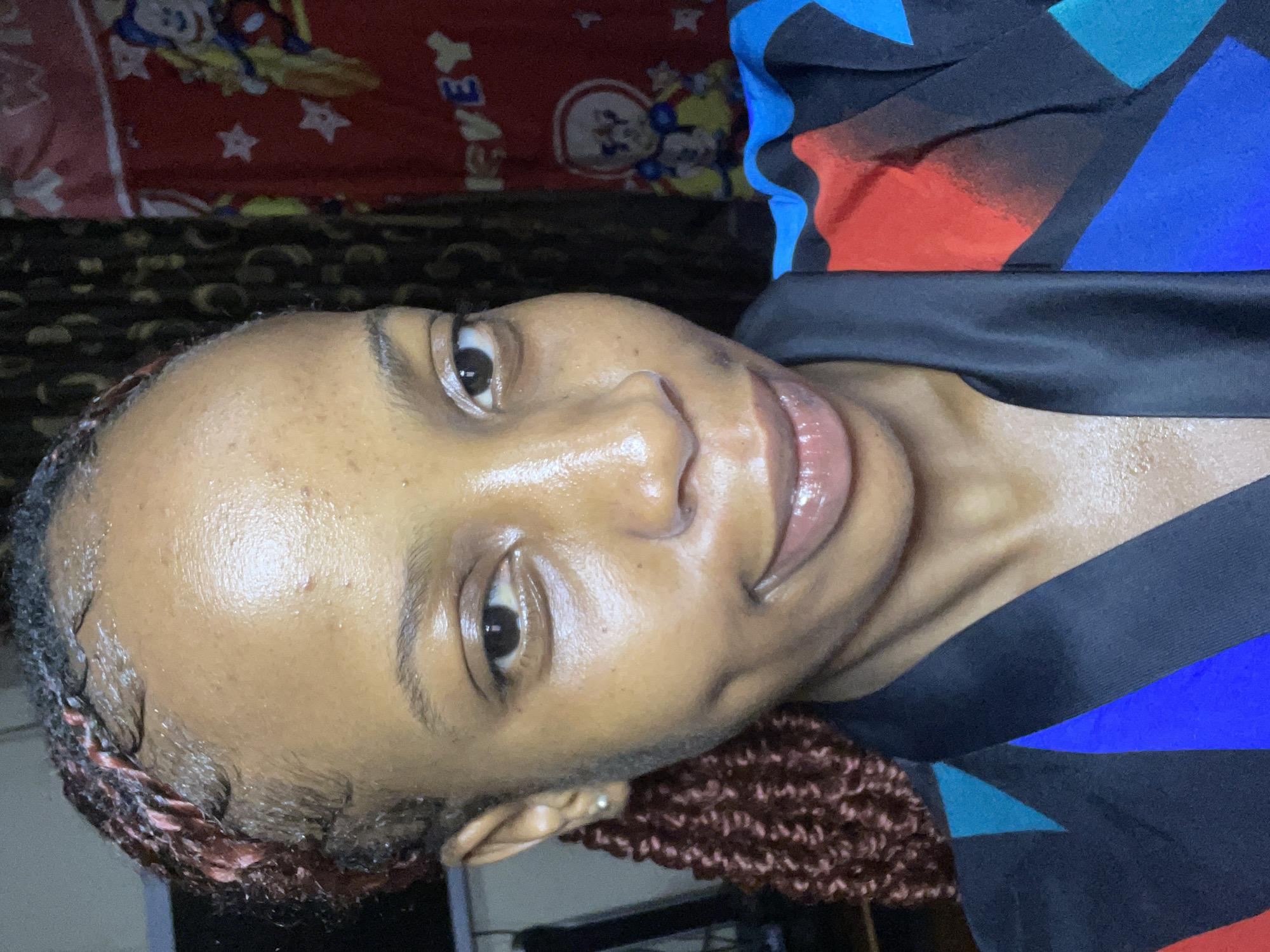 The image is a close-up selfie of a young Black woman with braided hair and dimples. She is wearing a colorful, abstract-patterned garment. The background appears to be indoors, with a dark curtain and a red fabric with images of cartoon characters, possibly a bedsheet or tapestry. The lighting suggests it is an indoor setting, and the overall impression is casual. There is no information in the image that indicates the location is the city of Jos, Nigeria.