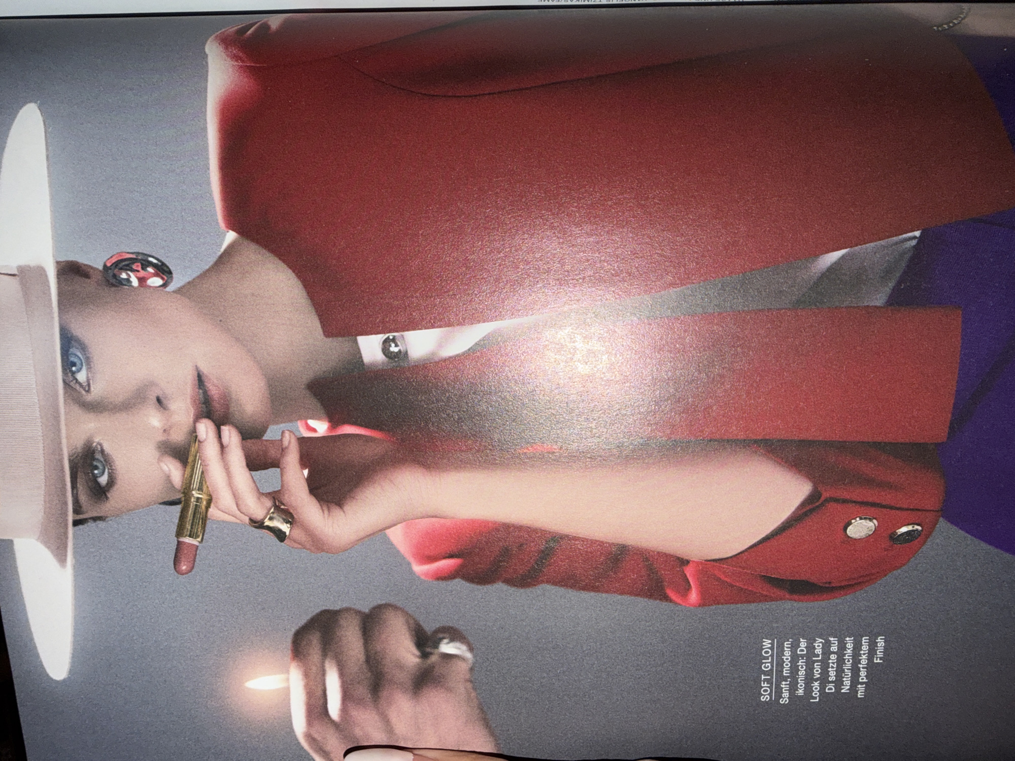 This is a studio photograph featuring a close-up of a person, appearing to be a woman, with striking blue eyes and fair skin. She is wearing a red blazer and a wide-brimmed hat. Her left hand is holding a lipstick, which she appears to be applying to her lips. A gold ring is visible on her right hand. The background is a plain, textured gray. The lighting is dramatic, creating strong highlights and shadows, suggesting a professional photo shoot.
On the right side of the image, there is text in a white font against a dark background, oriented vertically. The text reads: "SOFT GLOW Sanft, modern, ikonisch: Der Look von Lady Di setzte auf Natürlichkeit mit perfektem Finish." This translates from German to "Soft Glow Gentle, modern, iconic: The look of Lady Di relied on naturalness with a perfect finish." The overall aesthetic is one of glamour and beauty. The subject's pose and the focus on the makeup application suggest a theme related to fashion or cosmetics. The image is cropped to emphasize the face, hands, and the product.
