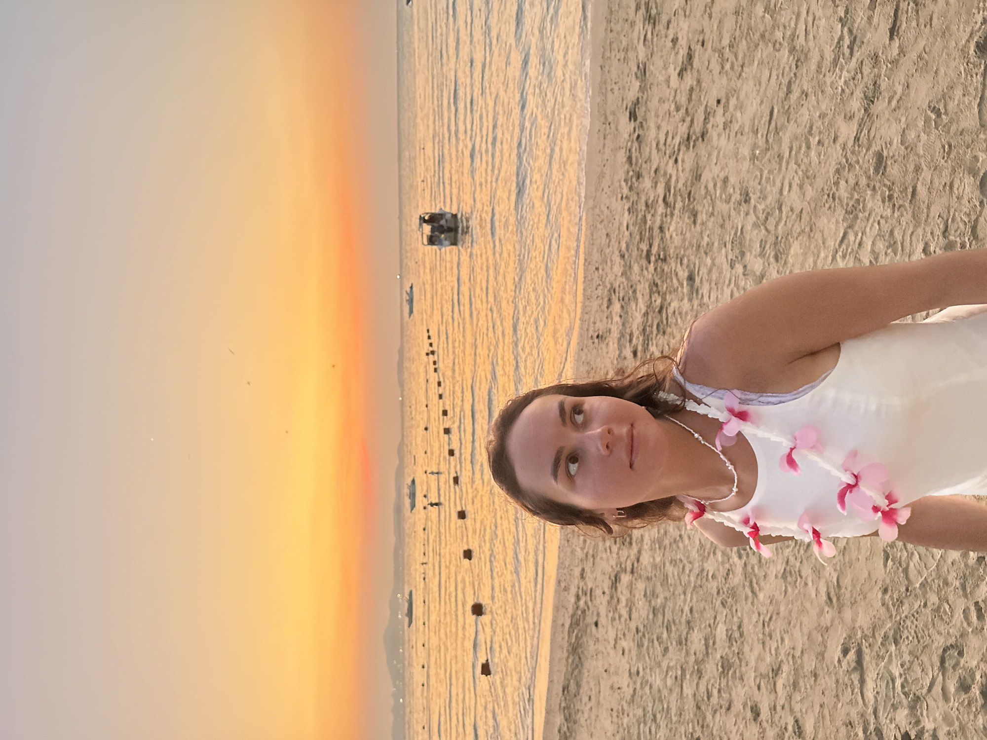 A woman wearing a white top and a pink flower lei stands on a beach in Pattaya City, Thailand. The beach is sandy, and the water reflects the sunset sky, which is orange and yellow. There are buoys in the water, and a vehicle is visible in the distance.