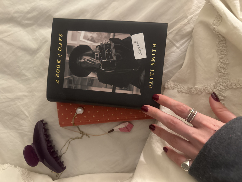 The image displays a flat lay composition of personal items on a white, textured, rumpled fabric surface, consistent with bedding, in an indoor setting. The specific location within New York City, United States of America, is not discernible from the provided visual information.
In the upper left, a dark purple hair claw clip is present. A gold-toned necklace, featuring a baroque pearl pendant and a pink and white tassel-like charm, is draped over an orange book with white polka dots, which is partially visible underneath. The central focus is a black hardcover book titled "A BOOK OF DAYS" by "PATTI SMITH." Its cover displays a black and white photograph of a person wearing a hat and holding a camera. A small white rectangular sticker with the handwritten word "signed" is affixed to the lower right portion of the photograph on the book cover.
A human left hand is positioned in the lower-left quadrant. The fingernails are painted with dark red polish. A plain silver-toned ring is worn on the ring finger, and multiple stacked silver-toned rings are on the middle finger. The wrist and lower arm are covered by a grey knitted sleeve. The hand rests gently on the white fabric surface, near the books. The overall scene suggests a quiet, domestic environment.