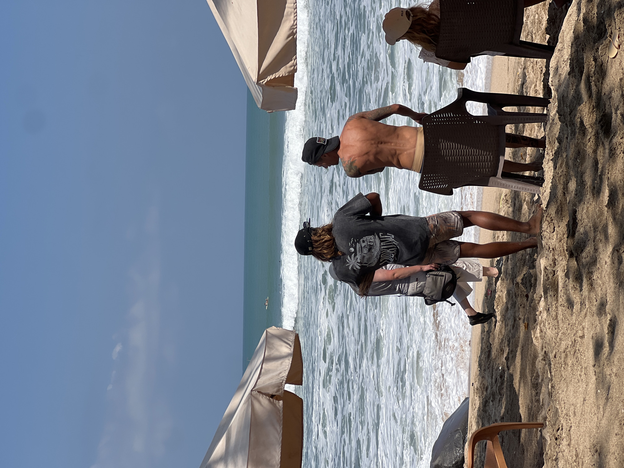 In Kuta, Indonesia, on a sandy beach, two men and a person are standing near the water's edge. One man, facing away from the camera, wears a black t-shirt with a graphic design and patterned shorts. Another man, also facing away, is shirtless and wears a backward baseball cap and beige shorts. A third individual, partially visible, walks away from the camera, wearing a grey t-shirt, beige shorts, and carrying a grey bag over their shoulder. In the foreground, two chairs are positioned on the sand. One chair is occupied by a person wearing a light-colored hat and reading. The ocean, with moderate waves, stretches out towards the horizon under a clear blue sky. Two large beach umbrellas are visible on either side of the frame.