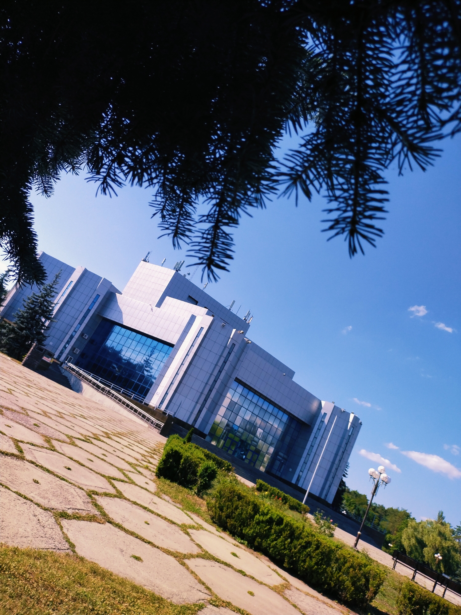 The image shows a modern, multi-story building with a glass facade and white exterior panels. The building appears to be an office or institutional structure. In the foreground, a paved walkway made of large, irregularly shaped stones is visible, with patches of grass growing between the stones. Lush green hedges line the walkway and the base of the building. A street lamp with three globes stands on the right side of the image, and other, smaller lamps are visible further in the distance. Evergreen trees are planted near the building and along the pathway.
The scene is viewed from a low angle, with the branches of a dark evergreen tree silhouetted against a clear blue sky in the upper portion of the frame. A few wispy white clouds are scattered across the sky. The sunlight suggests it is daytime, likely late morning or early afternoon, with bright illumination and clear visibility.
The image was taken in Horishni Plavni, Ukraine. There is no visible text in the image. The overall impression is one of a well-maintained public space or corporate campus on a pleasant day. No people or specific activities are depicted, focusing instead on the architecture and surrounding landscape.