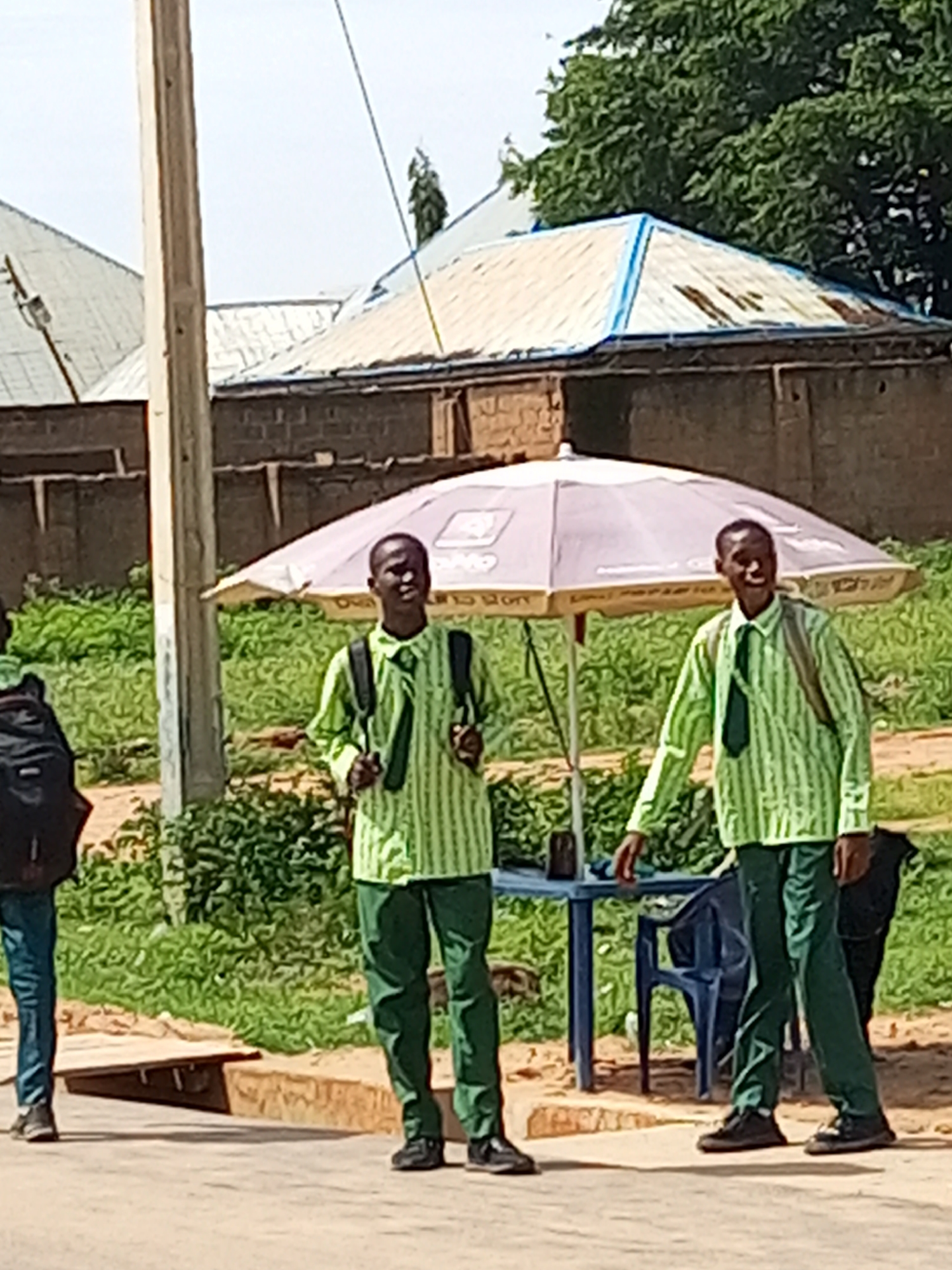 Two young boys in light green striped uniforms with dark green ties and trousers stand under a large, light-colored umbrella. They are both wearing backpacks. The boy on the right is looking towards the camera with a slight smile, while the boy on the left is facing slightly away. Behind them, a blue plastic table and chair are visible. The scene appears to be outdoors, with lush green foliage and a building with a corrugated metal roof in the background. A utility pole stands to the left of the umbrella. The ground is a mix of dirt and pavement. The lighting suggests it is daytime, likely midday, with the sun casting shadows. The overall mood is casual and ordinary, possibly a school break or waiting for transport. The image lacks any visible text. The setting appears to be Tempure, Nigeria, given the environmental context.