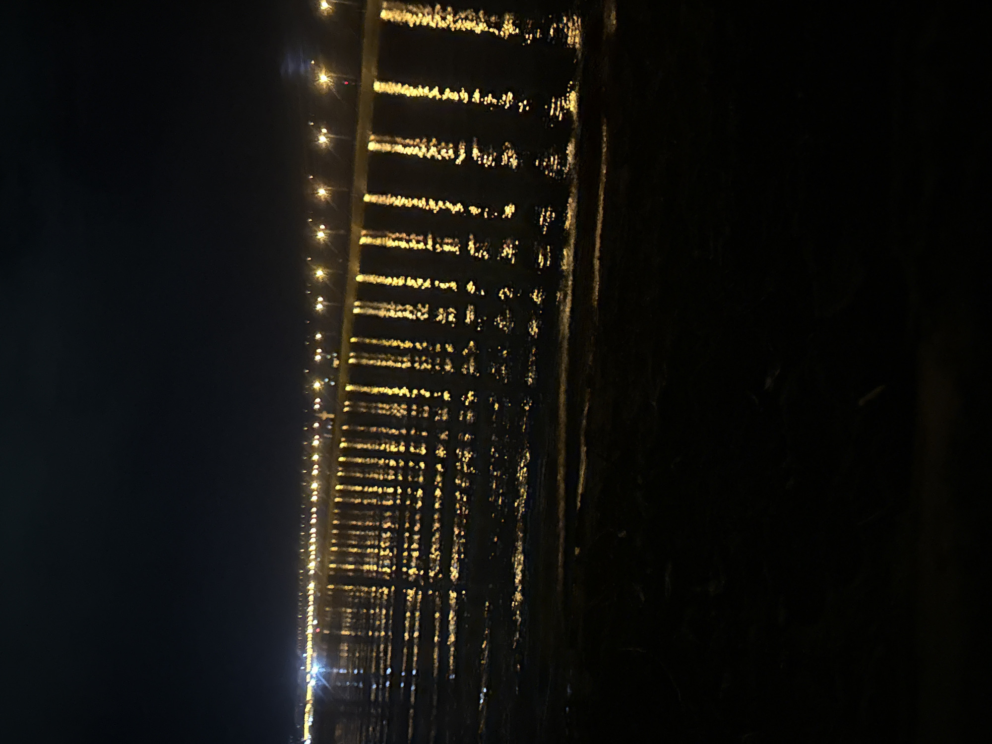 The image depicts a nighttime scene along a waterfront in Noviy, Russia, likely a promenade or embankment. A long line of streetlights illuminates the area, casting reflections on the water's surface. The water appears to be slightly rippled, creating broken, elongated streaks of yellow light. The foreground consists of dark, indistinct ground, possibly a beach or path. The sky is a deep, dark blue, with no visible celestial bodies or clouds. The overall impression is one of a quiet, well-lit urban waterfront at night.