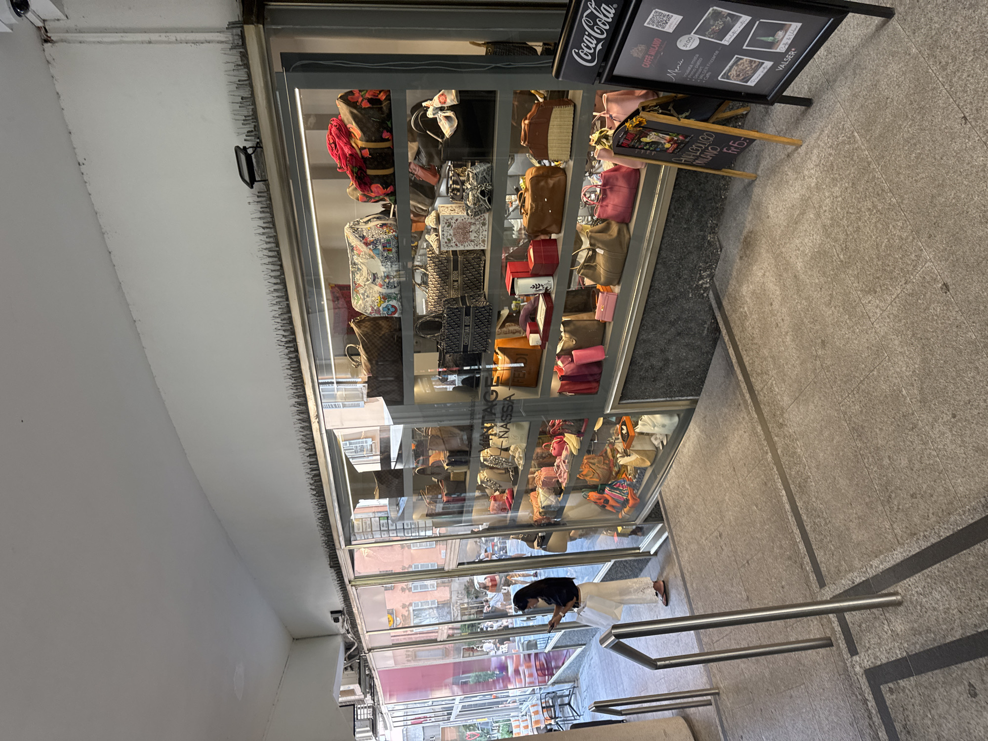 In Lugano, Switzerland, a street-level boutique displays a collection of high-end handbags and accessories behind a large glass storefront. The store's interior is visible, featuring multiple shelves packed with various bags, some with distinct designer patterns and colors. A woman, dressed in a white outfit and holding a white shopping bag, stands outside the store entrance, looking at her phone. Adjacent to the storefront, two chalkboards advertise offerings, one with the Coca-Cola logo and the other detailing menu items and prices, suggesting a nearby cafe or restaurant. The overall scene captures a typical urban retail environment with a focus on luxury goods.
