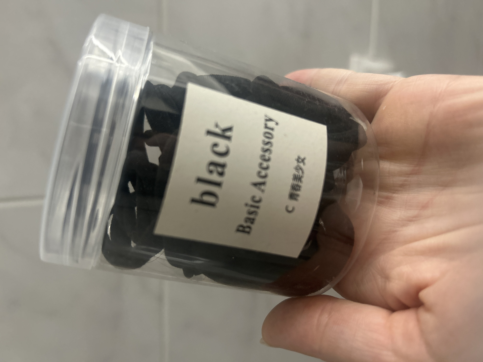 A clear plastic container filled with black hair ties is held by a person's hand. A label on the container reads "black Basic Accessory" in English, with Chinese characters below. The background appears to be a tiled wall. The image does not contain any information about the city of Нижние Муллы, Russia.
