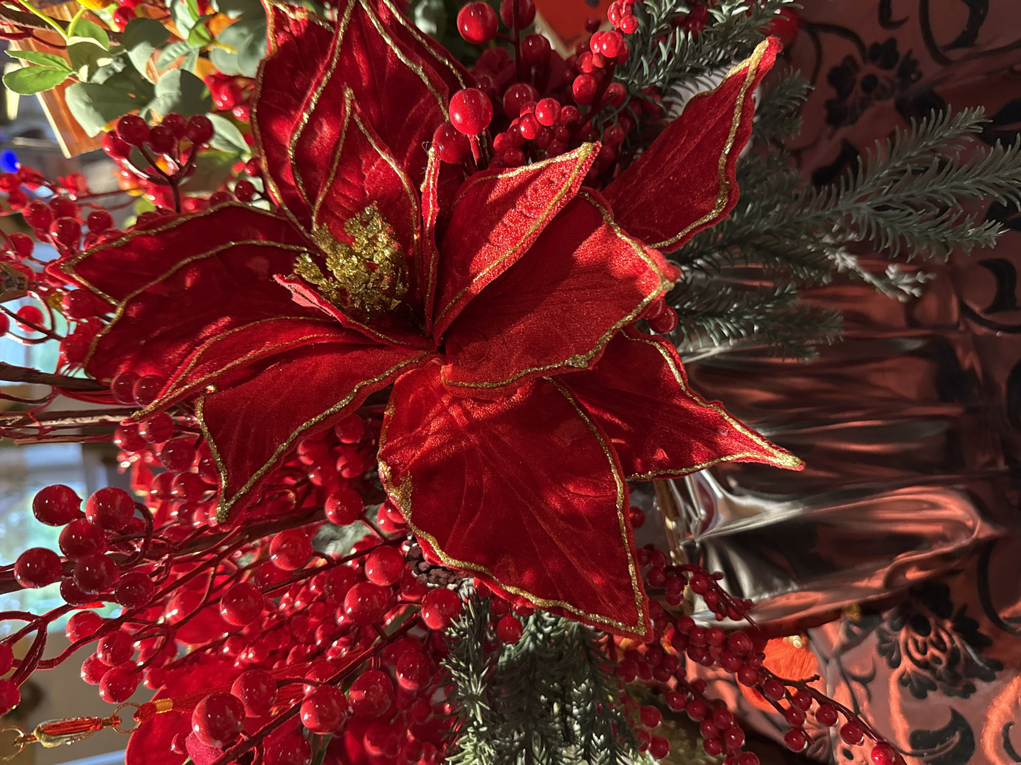 This is a close-up, eye-level, still shot of a festive holiday arrangement in Nordhausen, Germany. The arrangement is dominated by a large, red poinsettia with gold trim. Surrounding the poinsettia are numerous branches laden with small, bright red berries. Sprigs of artificial evergreen foliage are interspersed throughout the bouquet. The arrangement is displayed in a shiny, silver, pleated vase, which rests on a dark red tablecloth with an intricate black pattern. The lighting suggests an indoor setting, possibly a home or a shop, with a softly blurred background indicating ambient light. No people are visible in the frame.
