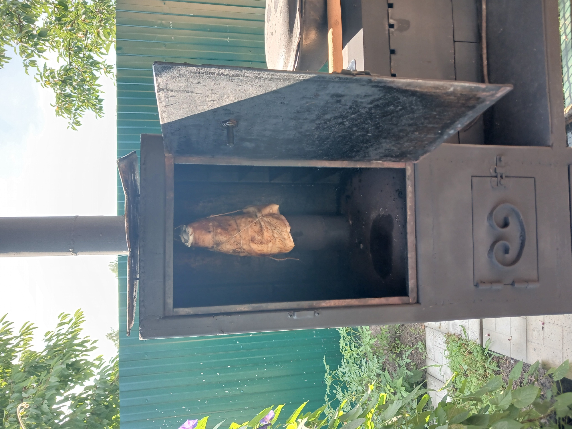 In a rural setting, possibly Maslova Pristan', Russia, an open, dark metal smoker stands in front of a green corrugated metal fence. A piece of meat, trussed with twine, hangs from the top of the smoker's interior. The smoker has a large, open door on the right side, revealing the dark interior. A small, decorative door with an ornate handle is visible on the lower front of the smoker. To the right of the smoker, a dark, round pot with a lid rests on a wooden surface. Lush green foliage, including a flowering vine and tomato plants, grows around the base of the smoker. The scene is illuminated by sunlight, casting shadows and highlighting the textures of the objects and vegetation. The sky above is a pale blue with scattered white clouds.