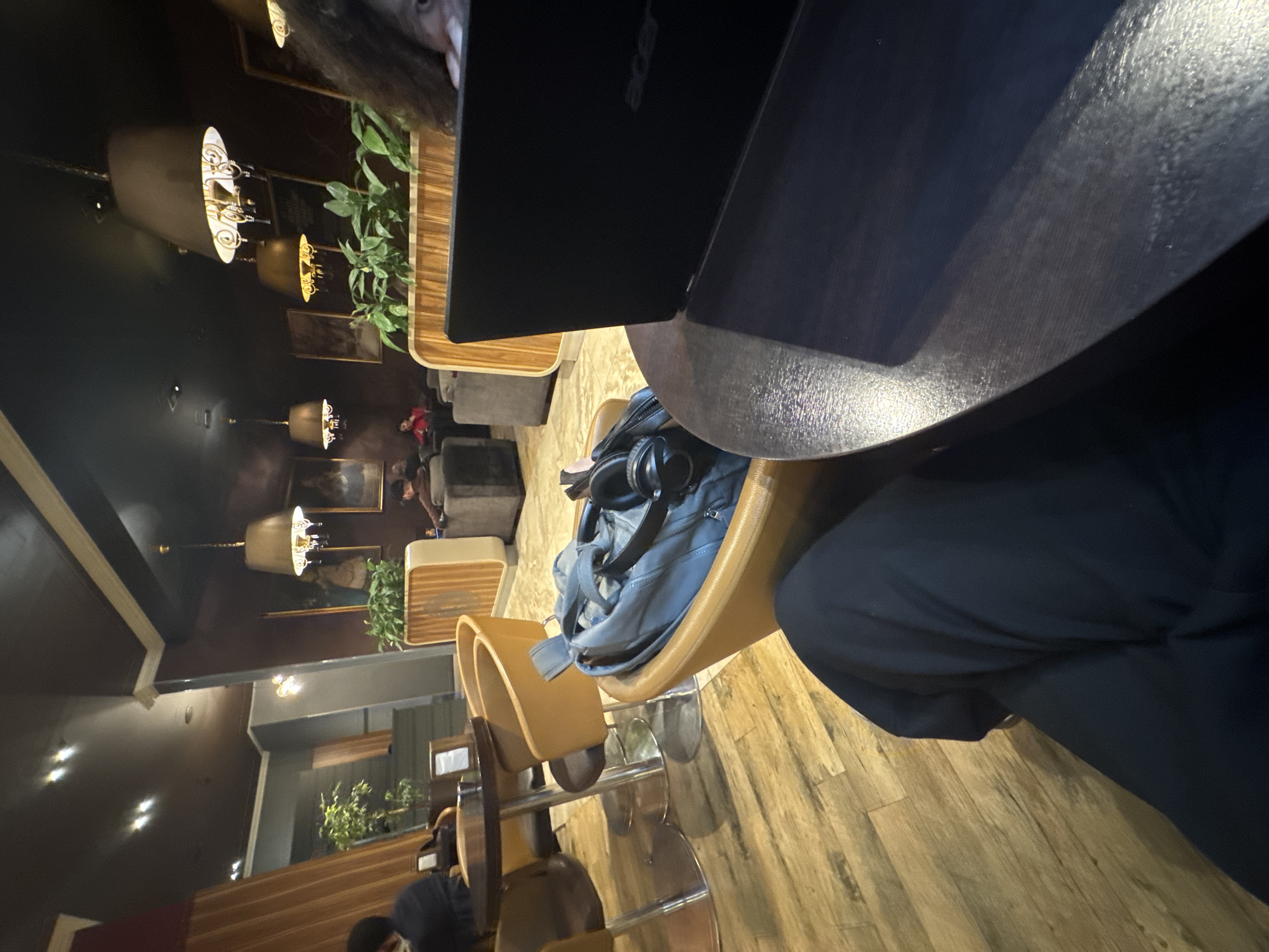 The image depicts the interior of a dimly lit commercial establishment, likely a cafe or lounge, located in Yerevan, Armenia. The scene is captured from a low vantage point, showcasing wooden plank flooring throughout.
In the immediate foreground, the dark-clothed legs and lap of an individual (the camera holder) are visible, positioned below a dark, round table. A black Acer laptop with its lid partially raised rests on this table. To the right of the laptop, a portion of a person's head with dark, curly hair is visible, facing towards the right side of the frame. Adjacent to the camera holder, a light brown, bucket-style armchair holds a light grey backpack with black over-ear headphones draped over it.
Moving into the midground, a second round table with a metallic base is visible to the left, surrounded by a dark brown chair and another light brown armchair. A person in dark attire is seated in the dark brown chair, facing away from the camera. Further back on the right, a wooden-slatted planter containing green foliage acts as a divider. Behind this planter, a person is seated in a dark grey upholstered armchair. Other dark grey armchairs and sofas are arranged throughout the space, defining several seating areas.
The background reveals dark brown or maroon walls adorned with framed artworks. Multiple ornate chandeliers with dark fabric shades and internal light sources hang from a dark ceiling, providing warm, ambient illumination, supplemented by recessed lights. Vertical ribbed wooden panels are integrated into the wall design. In the far background, another individual dressed in red clothing is visible, seated at a table. The overall environment suggests a sophisticated and relaxed indoor public space.