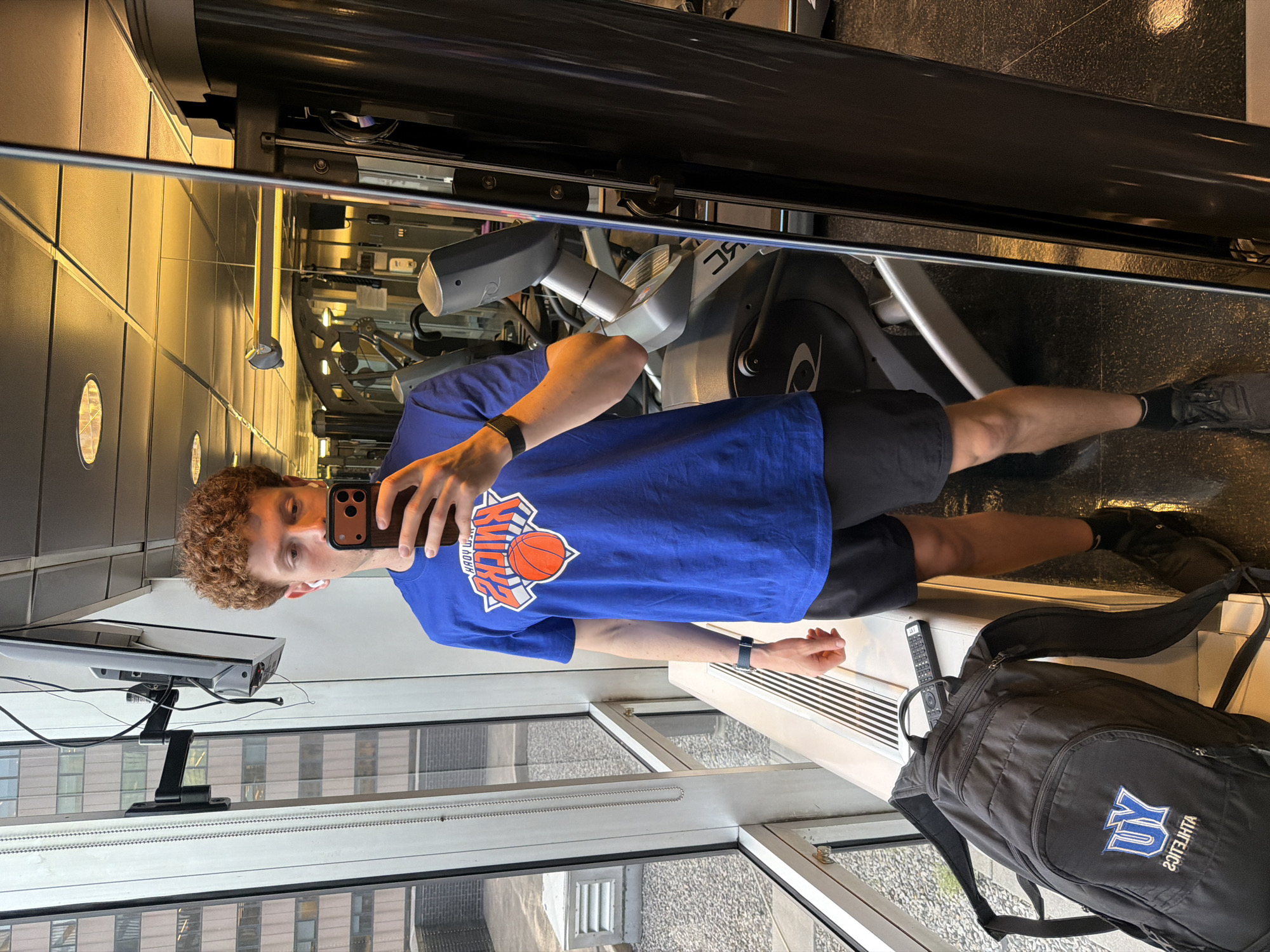 A young, white male with curly auburn hair, wearing a blue New York Knicks t-shirt and black shorts and sneakers, is taking a mirror selfie in a fitness center located high enough within a building to provide a view of other buildings in the city of New York, United States of America. Visible equipment includes a stationary elliptical trainer, a television, and a black backpack with the letter "U" on it is on the floor.