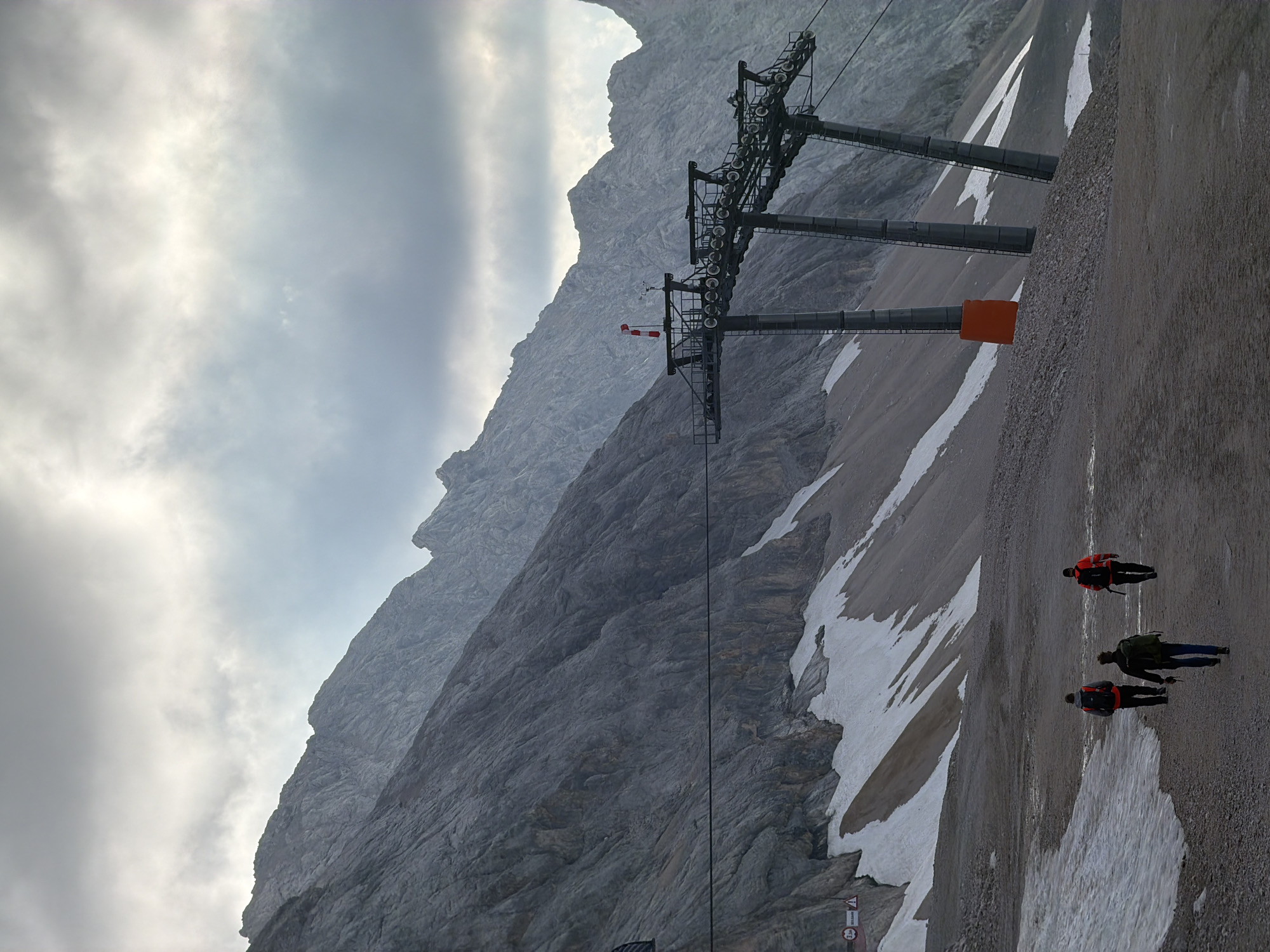 The image depicts a mountainous landscape, likely in the Alps, with a cable car station visible on the right. The mountains are rocky and steep, with patches of snow. In the foreground, three individuals are walking away from the camera on a gravel path. They are wearing backpacks and appear to be hikers. The sky is overcast with clouds. The scene suggests a remote, high-altitude location suitable for outdoor recreation. The presence of a cable car station implies this could be a popular tourist destination, possibly accessible by ski lift. While not explicitly stated, the general aesthetic suggests a European mountain range.