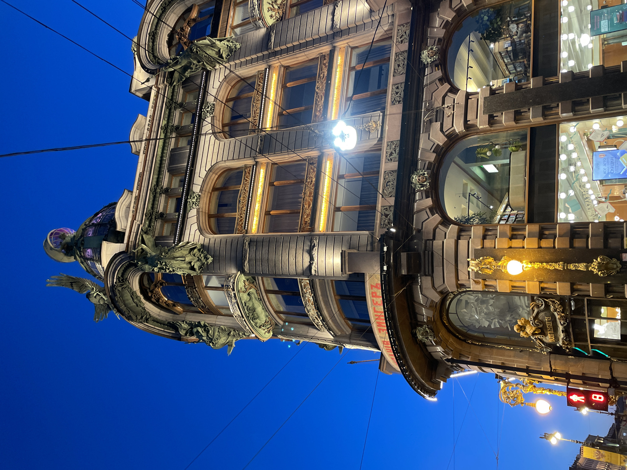 An elaborate, multi-story building, likely commercial, is depicted at dusk or night against a deep blue, clear sky. The building, viewed from a low angle and rotated in the original image, features a rich Art Nouveau or Eclectic architectural style. Its facade is adorned with numerous decorative elements, including ornate metalwork on balconies and window frames, intricate sculptures of winged figures, mythological creatures, and human faces that serve as corbels and decorative accents.
The building's windows are diverse: some are large arched display windows, while others are rectangular with decorative golden grates. Many windows are brightly lit from within, illuminating the interior and reflecting internal details like shelves and light fixtures. A distinctive glass dome, crowned with a sculpture, is visible at the building's corner.
Overhead wires, possibly for trams or electricity, crisscross the foreground against the dark sky. Streetlights with golden fixtures cast warm light onto the lower sections of the building. A traffic light showing a red "stop" signal for pedestrians and a partially visible "8" (possibly a timer) is also seen at the bottom right.
Visible text includes "КНИЖНАЯ" (Bookstore) and "ВСЕ ЛЕТО СКИДКИ" (All Summer Discounts) on display windows, and "В. НАЛИЧИИ" (In Stock/Available) near another window. Part of a red sign reads "З ДЕРЖИ..." (partially obscured). The building's grandeur and illuminated details suggest a prominent landmark in an urban setting, consistent with Saint Petersburg.