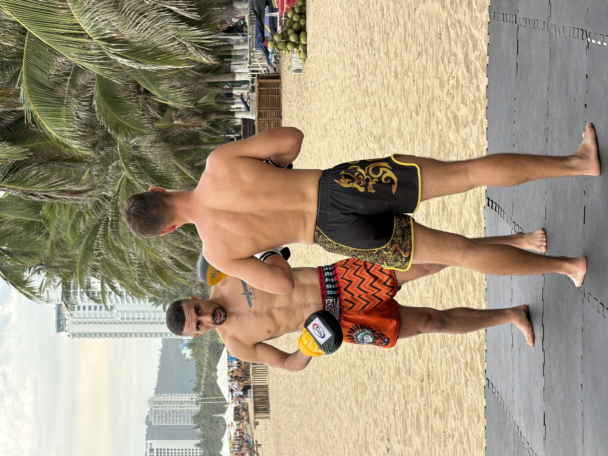 Two shirtless men are engaged in Muay Thai training on a sandy beach in Nha Trang, Vietnam. The man on the left, with a tattoo on his chest, wears orange Muay Thai shorts with a zigzag pattern and a yellow and black boxing glove on his left hand. He is facing the man on the right, who wears black Muay Thai shorts with a gold dragon design and also has a boxing glove on his left hand. They are standing on black mats. In the background, palm trees line the beach, with several tall buildings visible beyond the trees. Several white umbrellas and beach chairs are set up further down the beach, and a pile of coconuts is visible near some wooden structures. The sky is overcast.