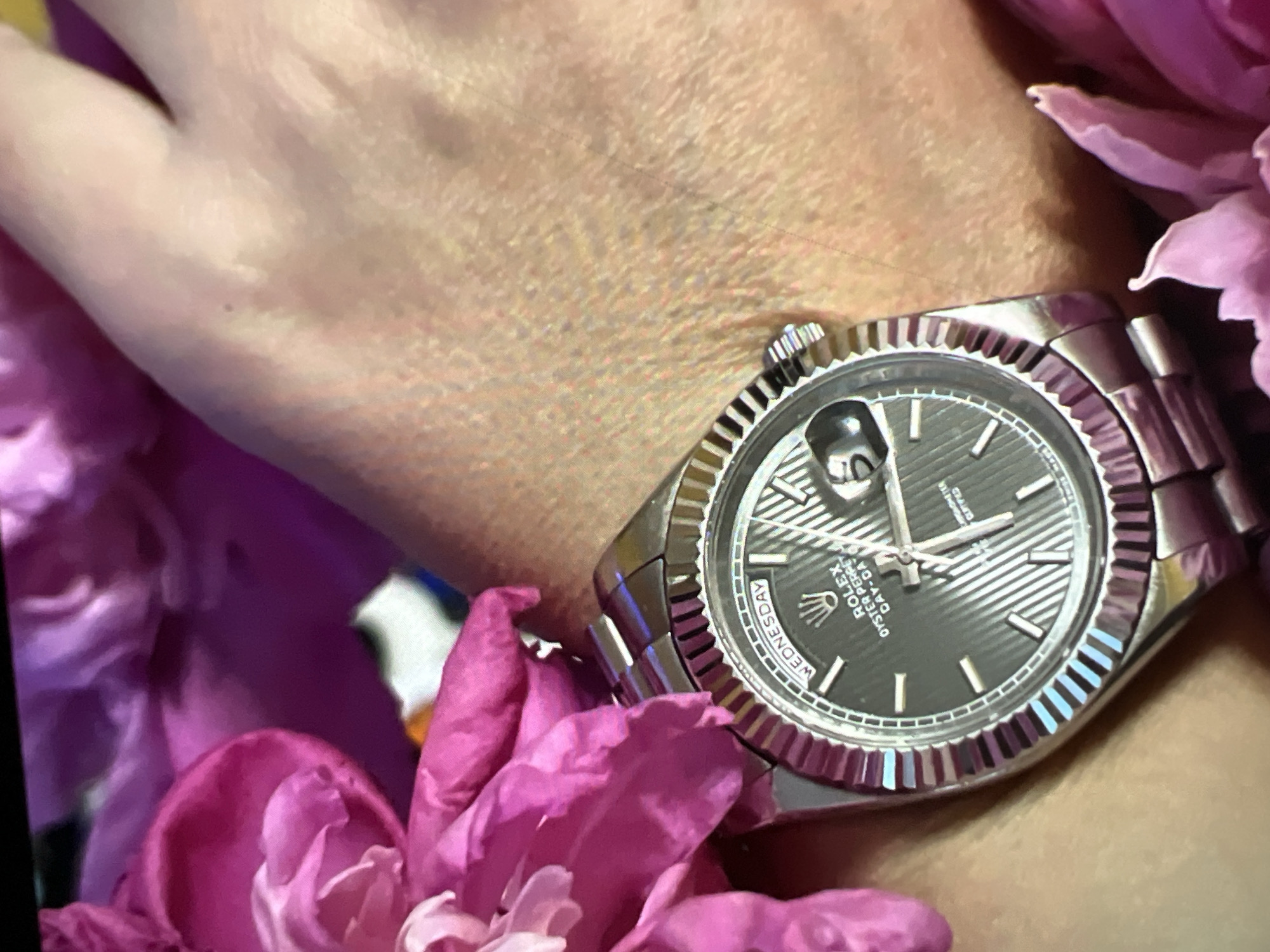 The image shows a close-up of a person's wrist wearing a Rolex Day-Date watch. The watch has a dark dial with silver hour markers and a fluted bezel. The day window at 12 o'clock displays "WEDNESDAY" in English. The watch is surrounded by pink and purple flowers. The skin tone is fair. The background is out of focus, but appears to be indoors or in a brightly lit area. There are no visible landmarks or contextual clues to suggest a specific location within the city of Minsk, Belarus.