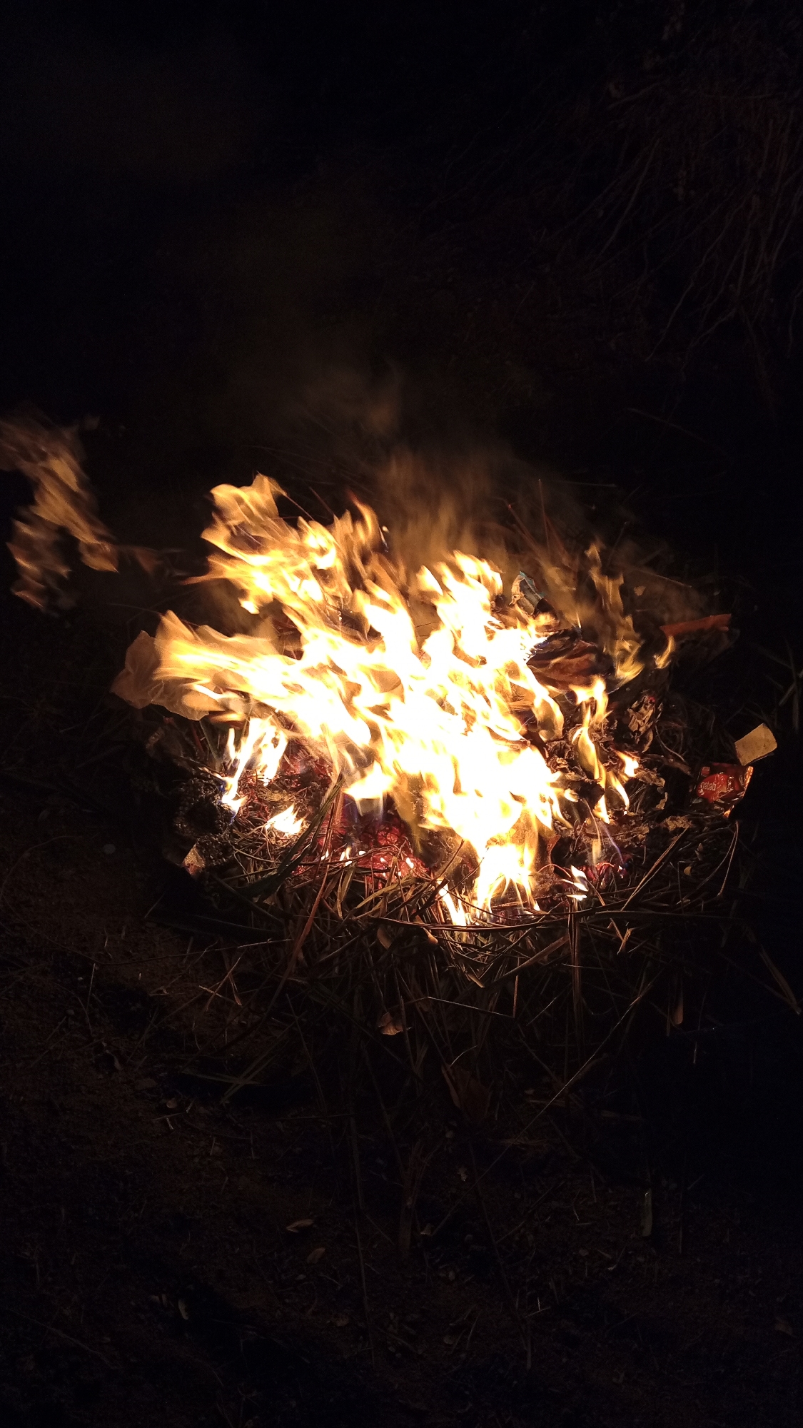 An outdoor scene at night captures a bonfire burning brightly with large flames. The fire is situated on dark ground, appearing to be a mix of soil and dry vegetation, such as twigs and leaves. Smoke rises from the fire, partially obscuring the dark, indistinct background which suggests trees or foliage. The flames are predominantly yellow and orange, illuminating the surrounding area. No people are visible in the image. The scene appears to be located in an undeveloped or natural area, as suggested by the presence of dry vegetation and the dark, unlit surroundings. Without further information or context related to the city of Buketteukuh, Indonesia, it is not possible to ascertain the specific location within that city.