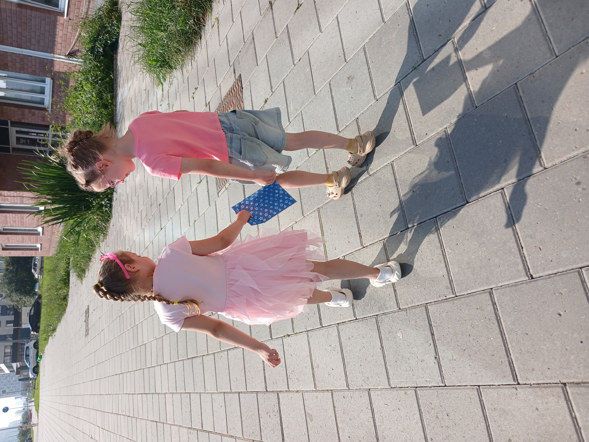 Two young girls walk on a paved sidewalk in front of a brick building. The girl on the left wears a pink dress and white shoes, holding a blue and white patterned object. The girl on the right wears a pink t-shirt, denim shorts, and beige clogs, looking down. The sidewalk is made of rectangular gray pavers. In the background, there is a building with windows, green grass, and a few parked cars. The scene appears to be in Yekaterinburg, Russia, based on the typical urban landscape.
