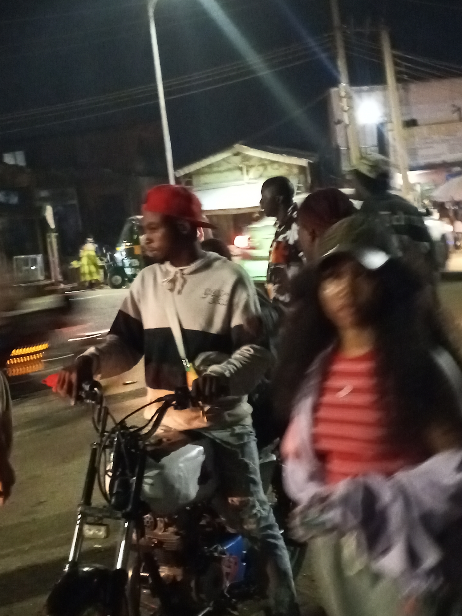A nighttime scene in Gwong, Nigeria, shows a man on a black motorcycle, wearing a red cap and a light-colored hoodie with a dark horizontal stripe. He is wearing blue jeans with rips in the knee area. A woman in a red striped shirt and a dark cap stands to his right. Other people are visible in the background. A street light illuminates the scene. There are buildings and other vehicles along the road. The overall atmosphere suggests a bustling city environment at night.