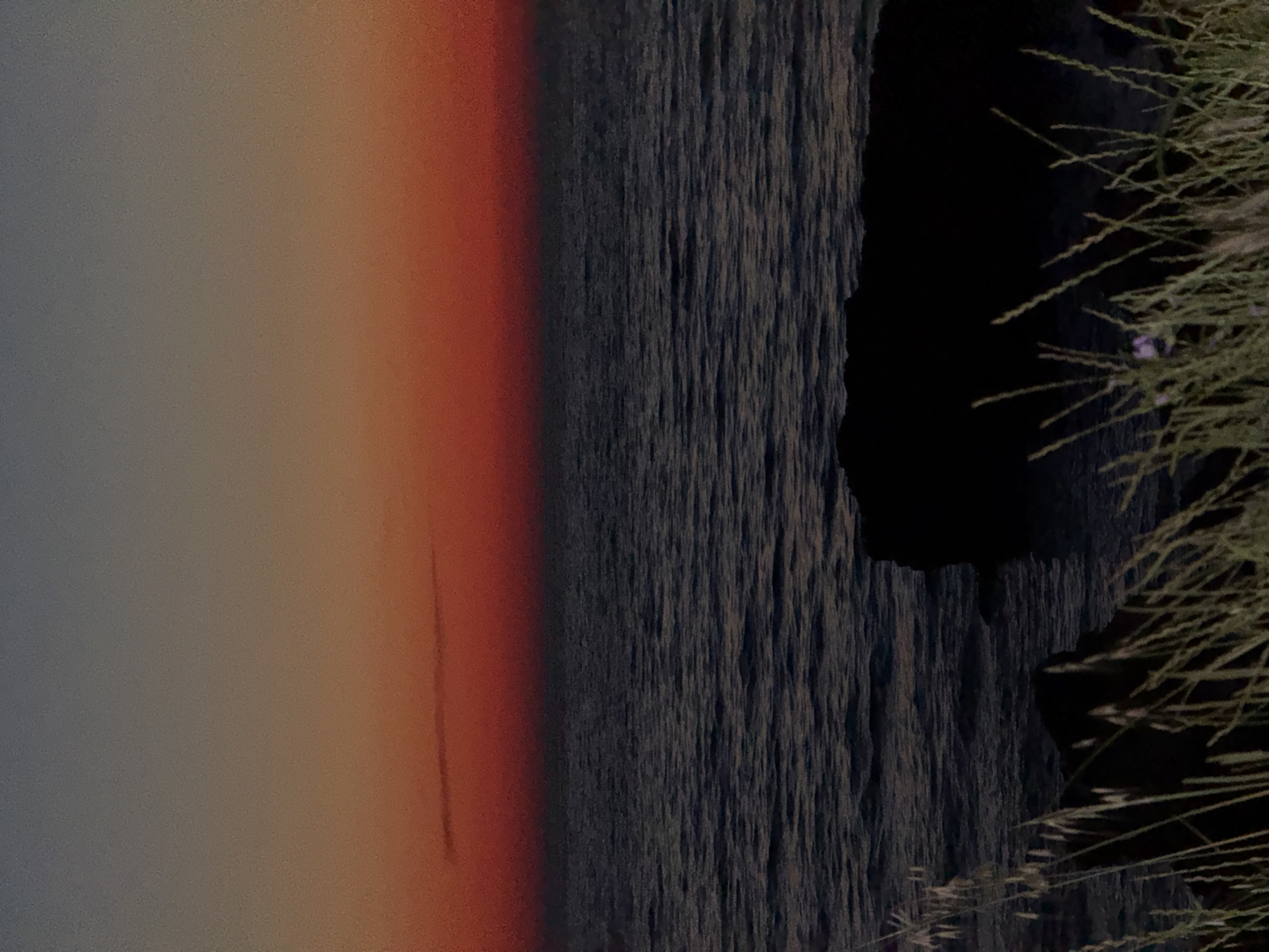 The image depicts a serene seascape at dusk. The sky is a gradient of colors, transitioning from a deep blue at the top to a vibrant orange and then a fiery red just above the horizon. A thin, elongated cloud is visible in the upper portion of the red band. The sea below is dark and choppy, reflecting the dim light of the sky. In the foreground, silhouetted rocks jut out from the water, partially obscured by tall, dry grass that sways gently. The overall impression is one of tranquility and natural beauty.