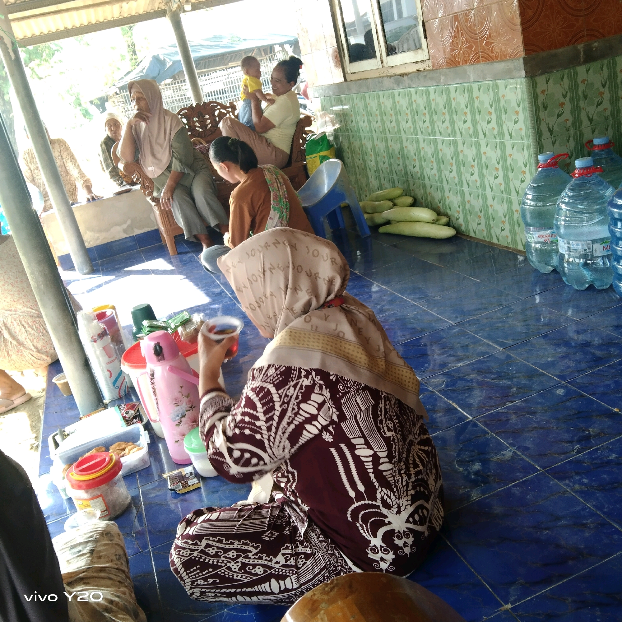 The image depicts an outdoor gathering of people, possibly in a communal space like a yard or under a shaded pavilion. The ground is tiled with large blue squares, some of which show wear and tear. In the foreground, a person wearing a maroon and white patterned garment and a beige headscarf is holding a small cup of dark liquid. Several items are scattered on the floor around them, including a pink thermos with floral patterns, a clear plastic container filled with what appears to be fried food, a red-lidded jar, and other miscellaneous objects.
In the mid-ground, several other individuals are visible. One woman, wearing a light-colored headscarf and gray pants, is seated on a decorative wooden chair, resting her chin on her hand. To her left, another person in a brown top and headscarf is seated. Further to the left, partially obscured, is a person in a patterned dress. In the background, a woman in a yellow shirt and beige pants is holding a baby. A blue plastic chair is also present, along with a stack of green elongated vegetables, possibly gourds or cucumbers, placed against a wall covered in patterned green tiles. Several large plastic bottles of water are lined up against the tiled wall as well. The overall atmosphere suggests a casual gathering, possibly a social event or mealtime.