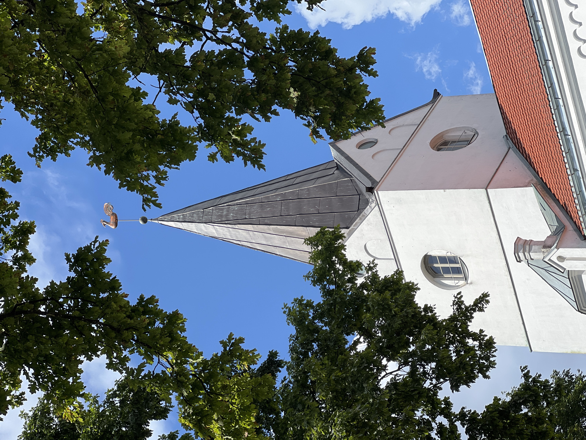The image shows the spire of a white church with a copper weather vane shaped like a rooster, against a bright blue sky with scattered white clouds. The spire is dark gray and conical. There are lush green tree branches framing the top of the image. Below the spire, a portion of the white church building is visible, featuring a round window and architectural details. A red-tiled roof of an adjacent building is also visible in the lower right corner. This is the Church of Jesus in Saldus, Latvia. No people are present.
