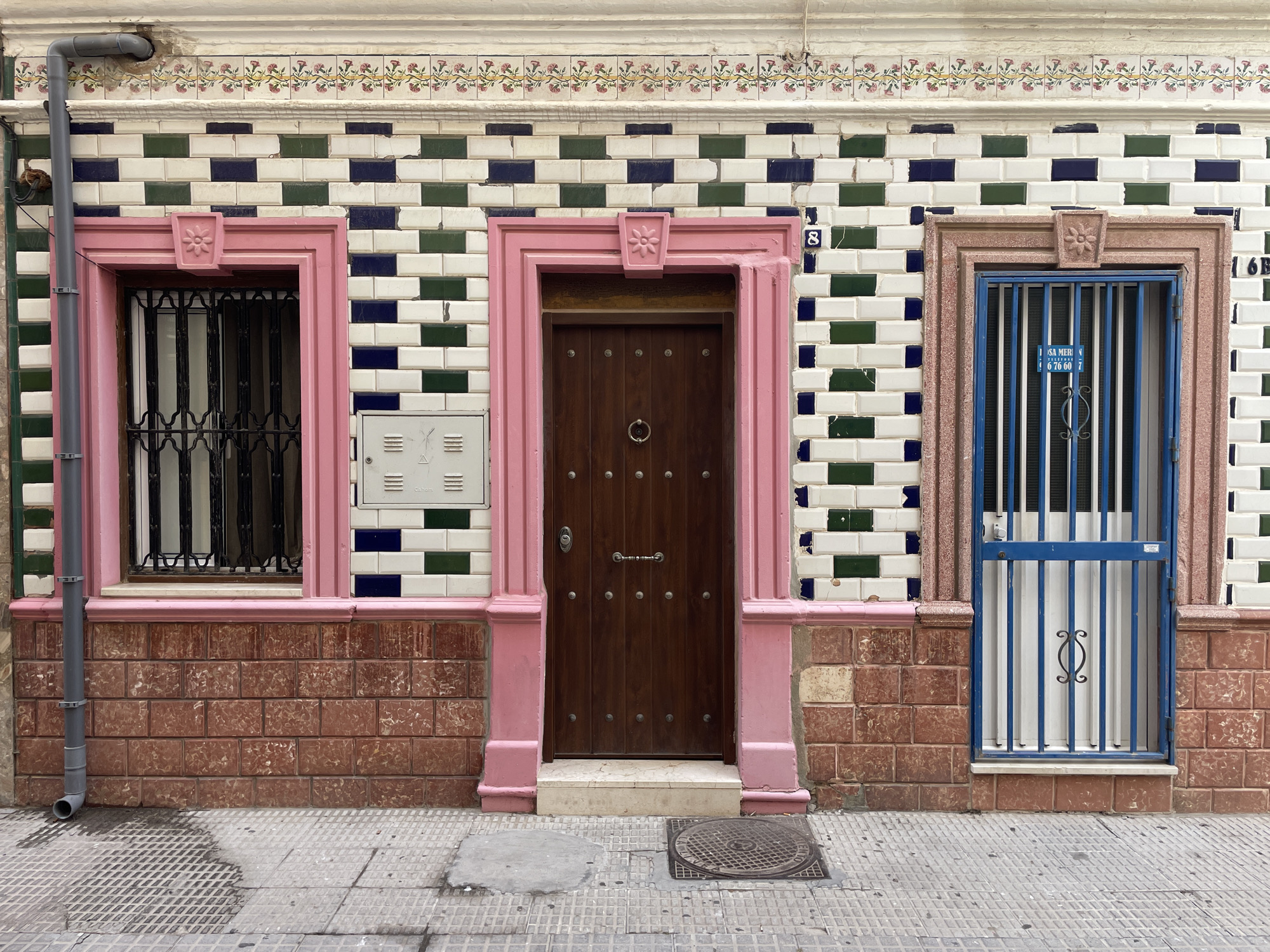 The image shows the exterior facade of a building in La Línea de la Concepción, Spain. The facade is adorned with a distinctive tile pattern, featuring alternating blocks of white, dark blue, and green tiles arranged in a staggered brickwork fashion. Above this tiled section, a decorative frieze with a floral motif is visible. The ground floor of the building is faced with reddish-brown textured tiles.
The facade features three openings. On the left is a window with a pink frame and a black wrought-iron security grate. To its right is a dark wooden door, also framed in pink, with a circular metal door knocker and several round studs. To the right of the door is a window with a blue metal grille and a white paneled door behind it. Both the window and the door are framed by the reddish-brown tiles.
A grey downspout runs vertically along the left side of the facade, adjacent to the window. A small, white electrical box is mounted on the wall to the right of the window. The sidewalk in front of the building is paved with grey square tiles, with a metal manhole cover and a circular patch of concrete visible. The overall scene is a street-level view of a building's entrance and window.