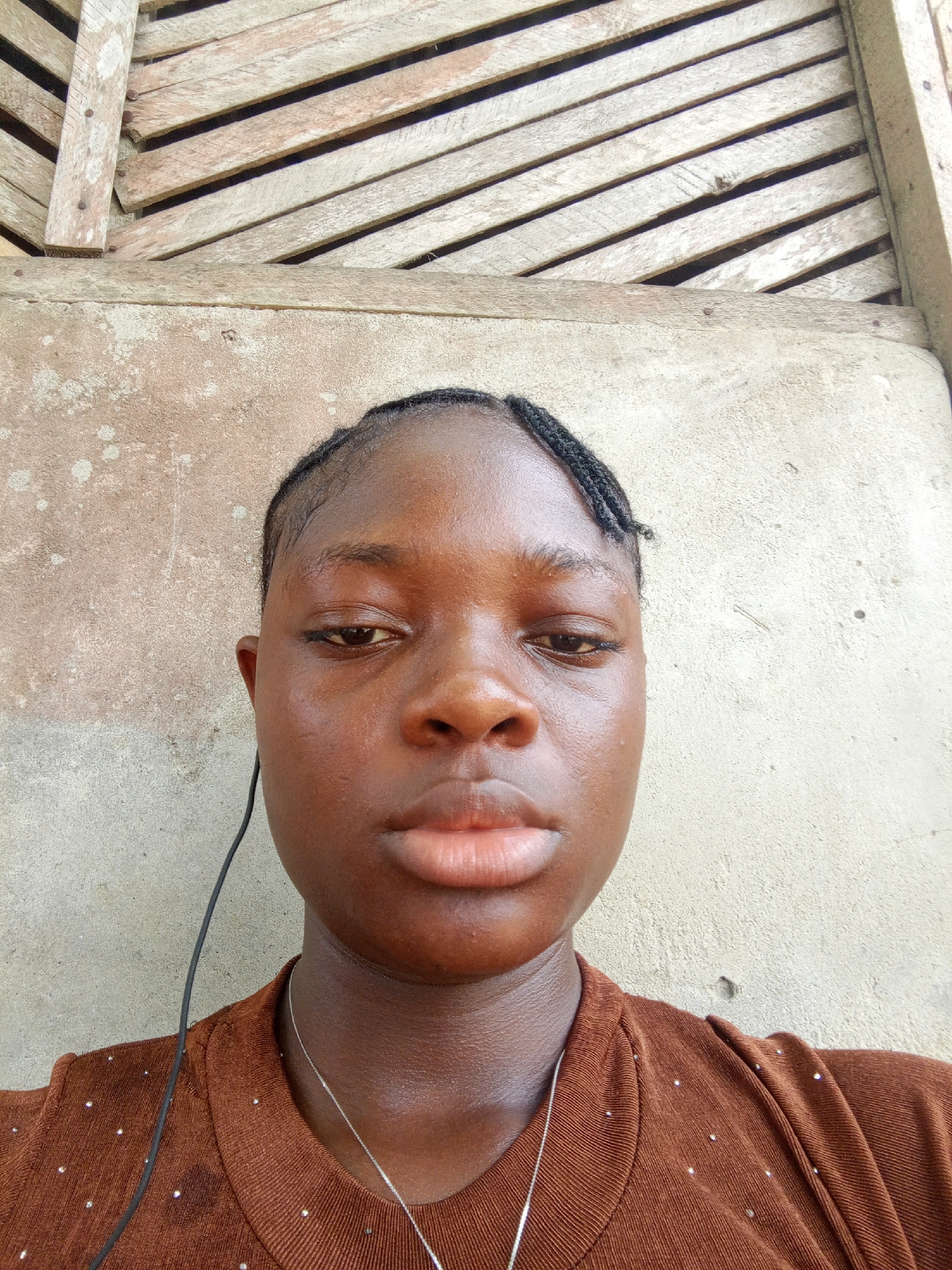 A close-up of a young Black woman is centered in the frame. She has dark skin, braided hair, and full lips. She wears a brown, collared shirt and a thin, silver necklace. A black wire dangles near her left shoulder. Behind her is a textured wall, partly obscured by a weathered, wooden slat structure. The location is in the city of Mbribit, Nigeria.