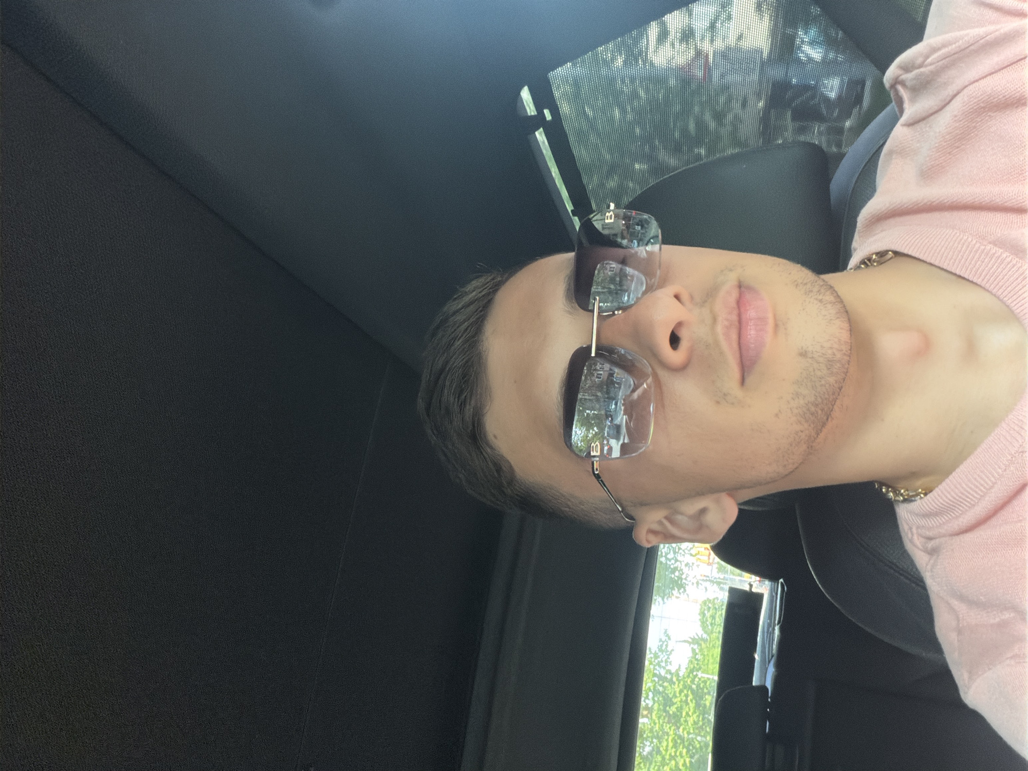 A medium-shot photograph shows a man wearing sunglasses and a pink long-sleeved shirt inside a vehicle. The vehicle's interior is visible, with black panels and a window showing trees and a partial view of a building. The man is wearing a gold chain. His face is angled towards the camera.