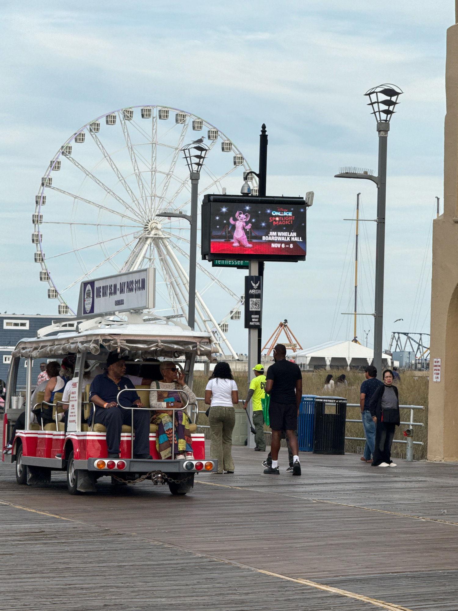 The image shows a bright, overcast day on the Atlantic City Boardwalk. A red and white tram carrying passengers is visible in the foreground. The tram is on a wooden boardwalk, with a large Ferris wheel in the background. A digital billboard displaying advertisements for "Disney On Ice Spotlight Magic!" is also visible. Other people are walking on the boardwalk, and there are various structures and signs that are typical of a boardwalk amusement area. The overall scene depicts a lively, public space in Atlantic City, New Jersey.