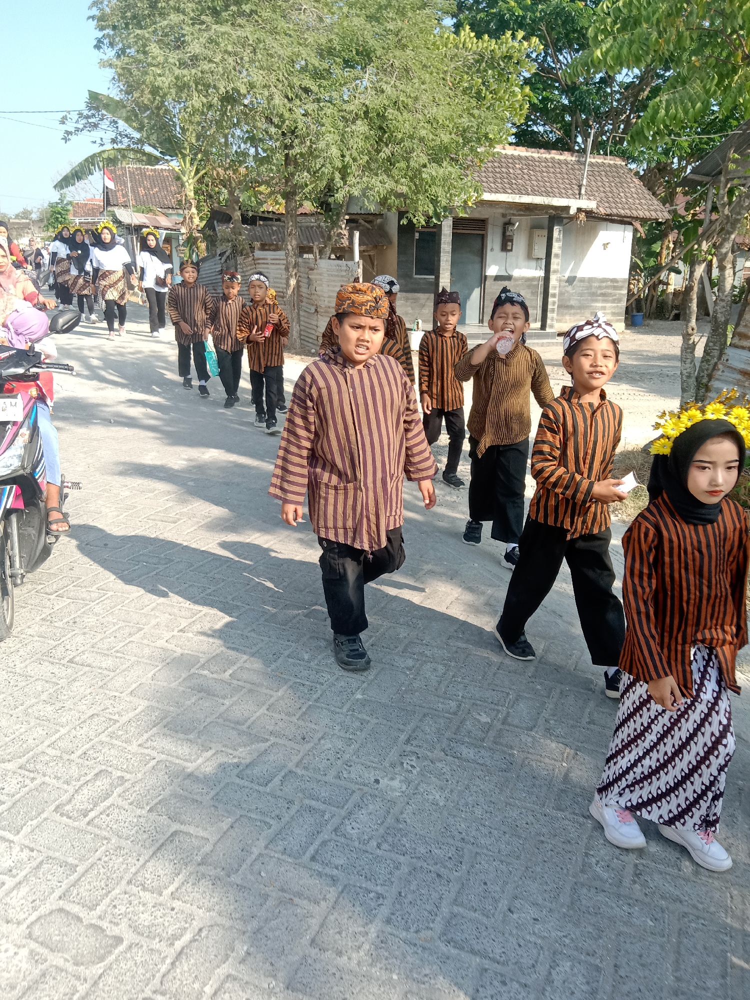 The image depicts a parade or procession on a paved street in Stiaji, Indonesia. A group of children, primarily boys, are walking in single file, dressed in traditional brown and white striped outfits and head coverings. One boy in the foreground is holding a small piece of paper. To the right of the boys, a young girl wearing a headscarf and a similar striped outfit is visible, adorned with yellow flowers. In the background, there are houses and trees, suggesting a residential area. A motorcycle is parked on the left side of the street. The scene is outdoors, under a clear sky.