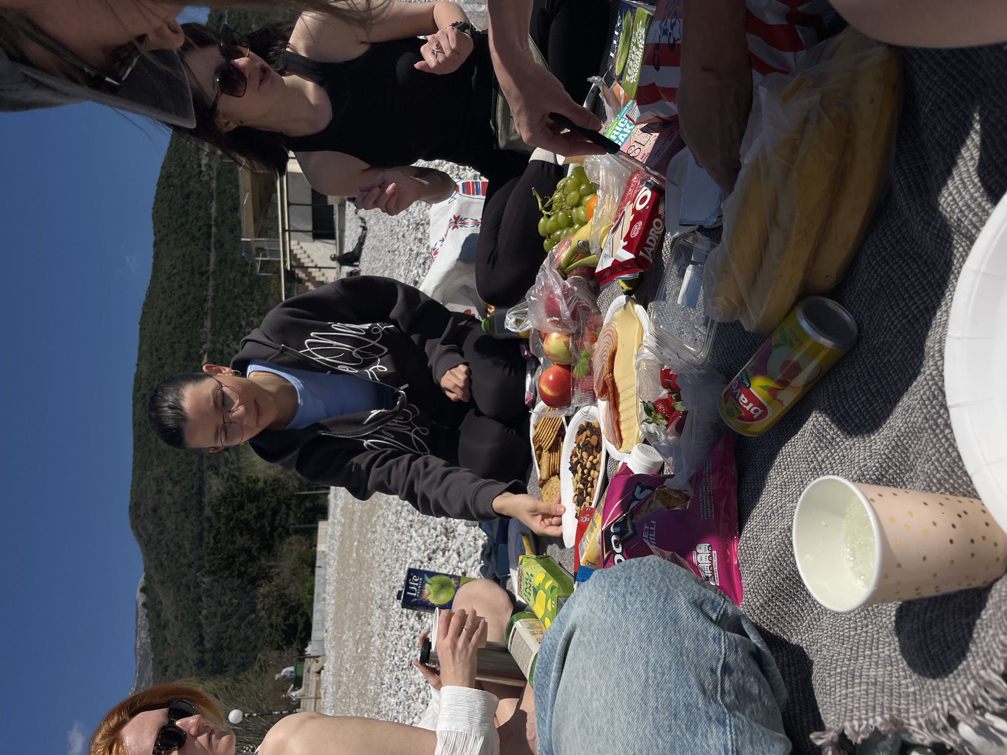 A group of people is having a picnic outside in Budva, Montenegro. The picnic is set up on a gray blanket on a rocky beach, with a view of a mountain and blue sky. Food items include grapes, bananas, apples, strawberries, cheese, salami, crackers, and mixed nuts. Beverages present are juice boxes and a drink in a pink cup. One person is wearing a black hooded sweatshirt and glasses, while another is wearing a white top and jeans. A woman in a black top and sunglasses is also part of the group. Some individuals are reaching for the food.
