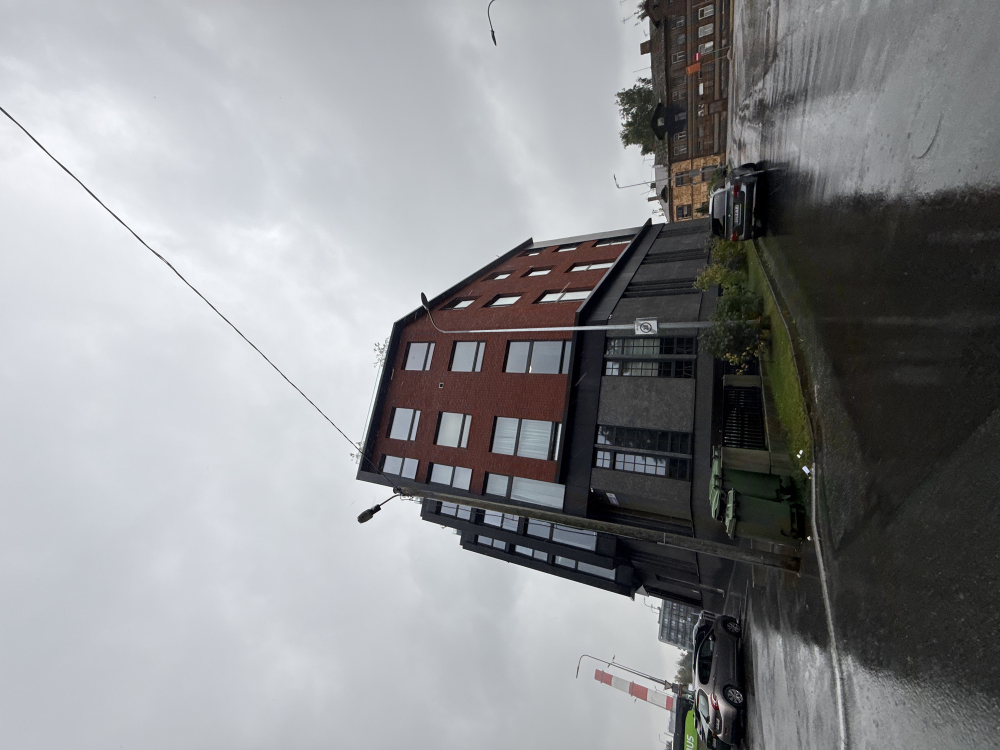 The image depicts a street scene in Tallinn, Estonia on an overcast day. A modern, multi-story building with a brick facade and dark lower section dominates the foreground, positioned at an angle to the camera. The building has numerous windows, some with curtains visible. Adjacent to the building are several green trash bins. A street lamp stands prominently in front of the building. A dark-colored SUV is parked on the wet asphalt road to the right of the building. Across the street, older brick buildings with multiple windows are visible. The road surface is wet, indicating recent rain, and reflects the overcast sky and surrounding structures. In the background, a tall structure with red and white segments, possibly a construction crane or industrial chimney, can be seen. A bus is partially visible on the left side of the image. The overall atmosphere is one of a damp, urban environment.