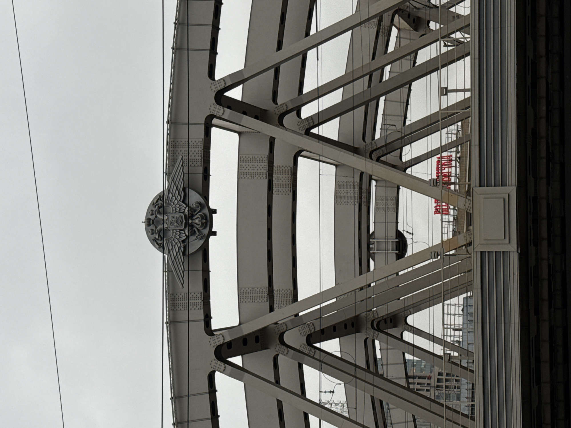 The image shows a close-up of the steel structure of the Rostokinsky Aqueduct in Moscow, Russia. The structure is painted in a light grey color. The image is taken at an upward angle, highlighting the bridge's arches, bracing, and riveted construction. A circular emblem with wings and a coat of arms is affixed to one of the main vertical supports. In the background, partially obscured by the bridge's framework, are some modern buildings, suggesting the aqueduct's location within an urban environment. A red sign with Russian lettering can be seen through one of the open spaces in the bridge.