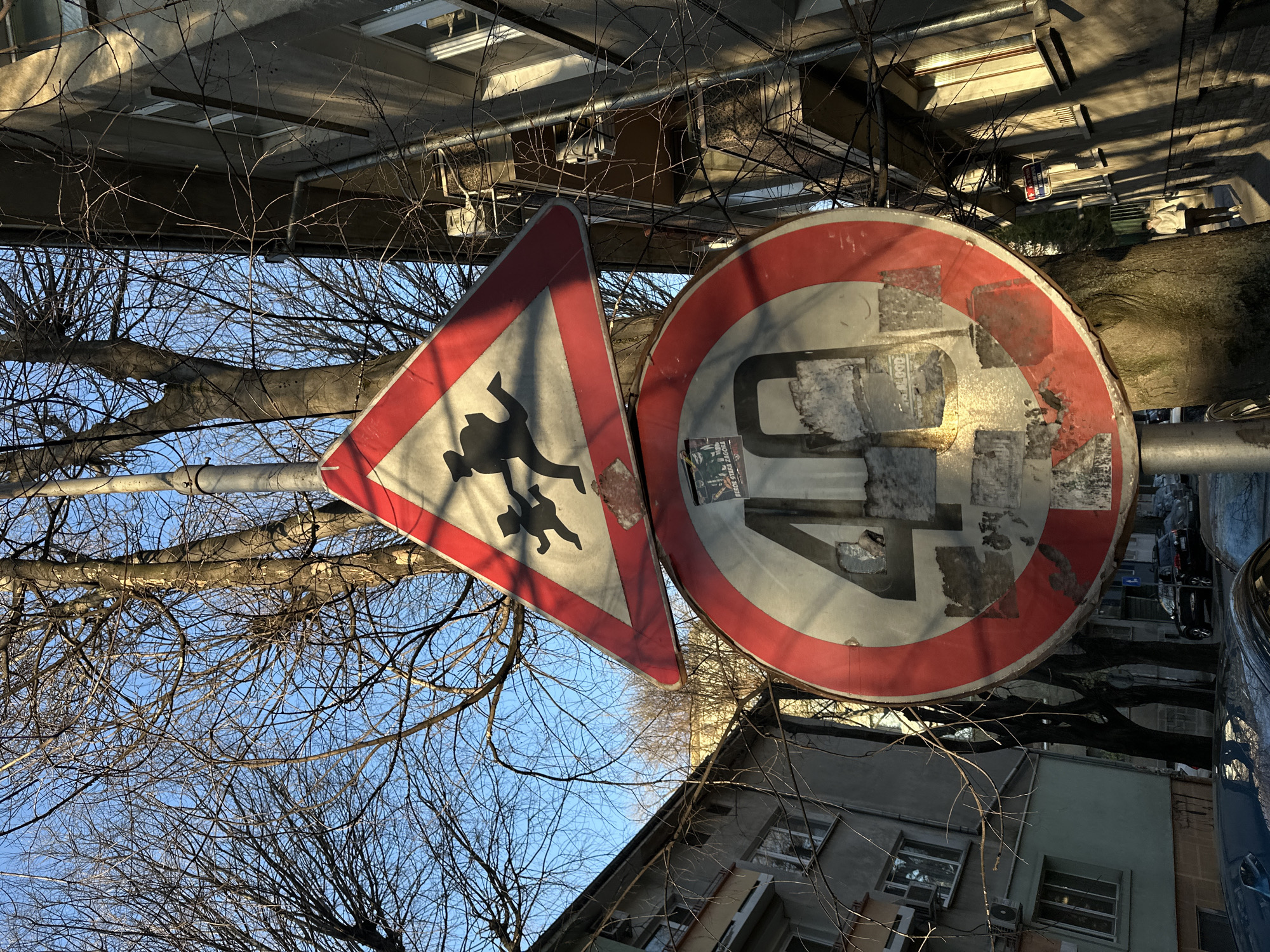 The image shows two road signs mounted on a pole. The sign on the left is triangular with a red border and depicts two silhouettes, presumably pedestrians. The right sign is circular with a red border indicating a speed limit of 40. Both signs are damaged. The background features buildings with windows and bare tree branches against a blue sky. The image appears to have been taken looking up at the signs. The location is in Stara Zagora, Bulgaria, as identified by visual elements characteristic of Bulgarian road signage.